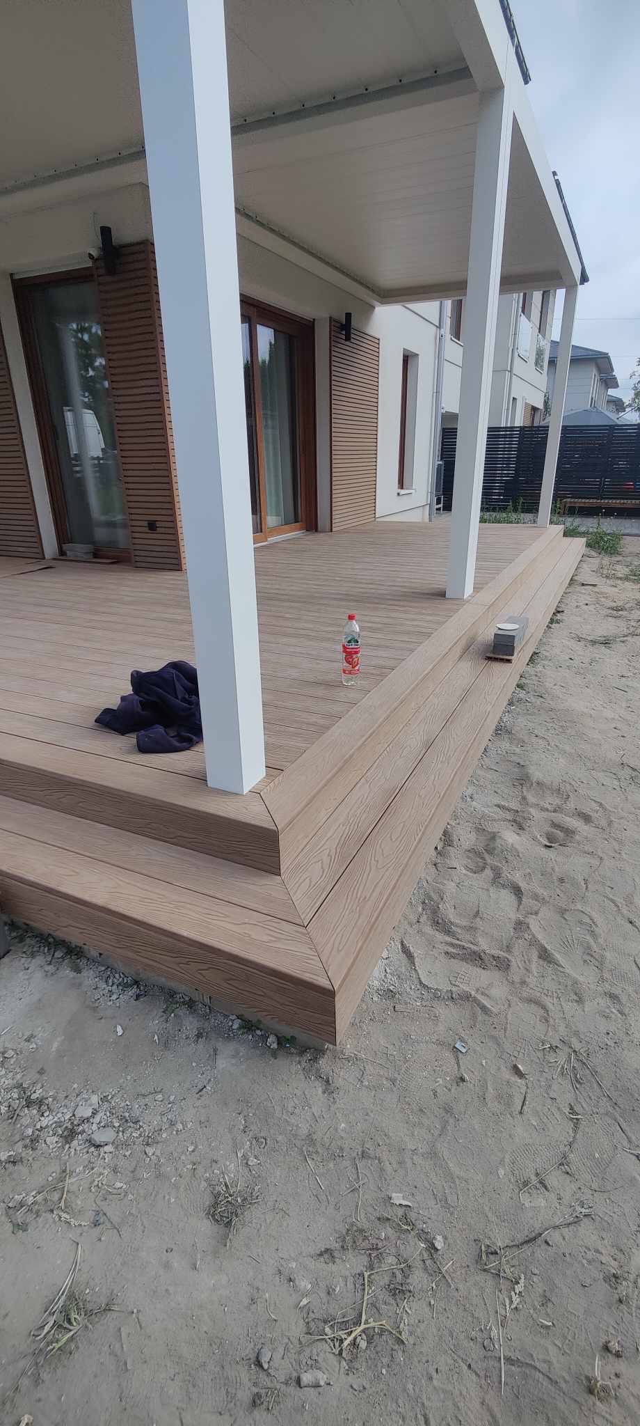 Nowoczesny taras z deski kompozytowej w odcieniach brązu, z białymi kolumnami podtrzymującymi zadaszenie. Na tarasie leży ciemna bluza i stoi butelka z napojem.