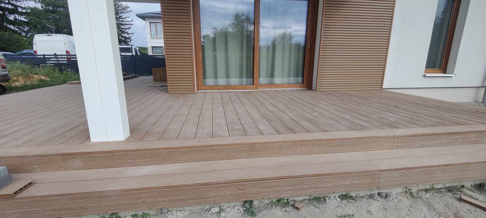 Nowoczesny taras z deski kompozytowej w odcieniach brązu, z widocznymi schodami i fragmentem elewacji budynku z drewnianymi panelami. Widok z bliska.