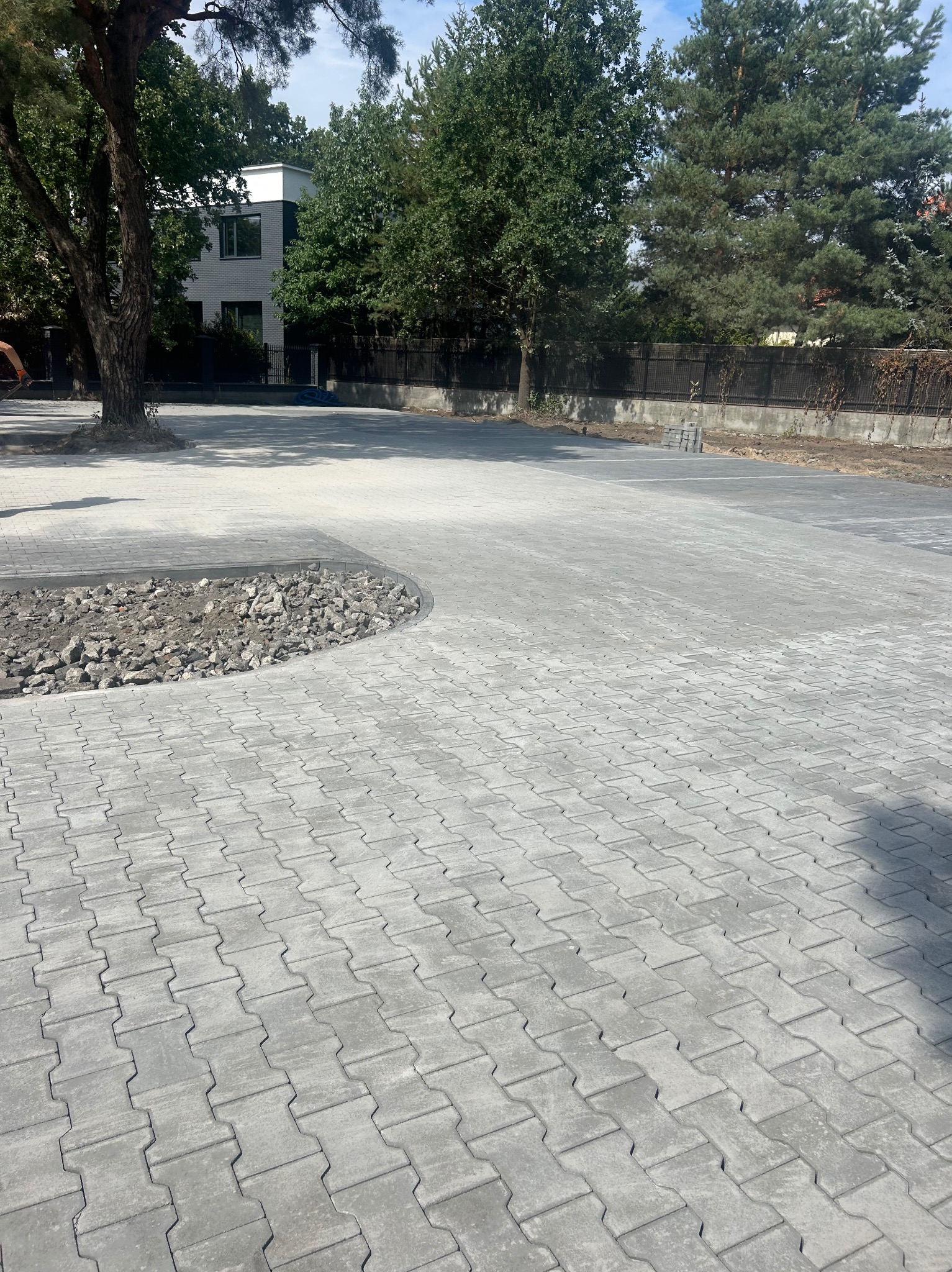 Szara kostka brukowa ułożona na dużej powierzchni, z obrzeżem wypełnionym kamieniami. W tle zieleń drzew i budynek. Widok z góry, słoneczny dzień.