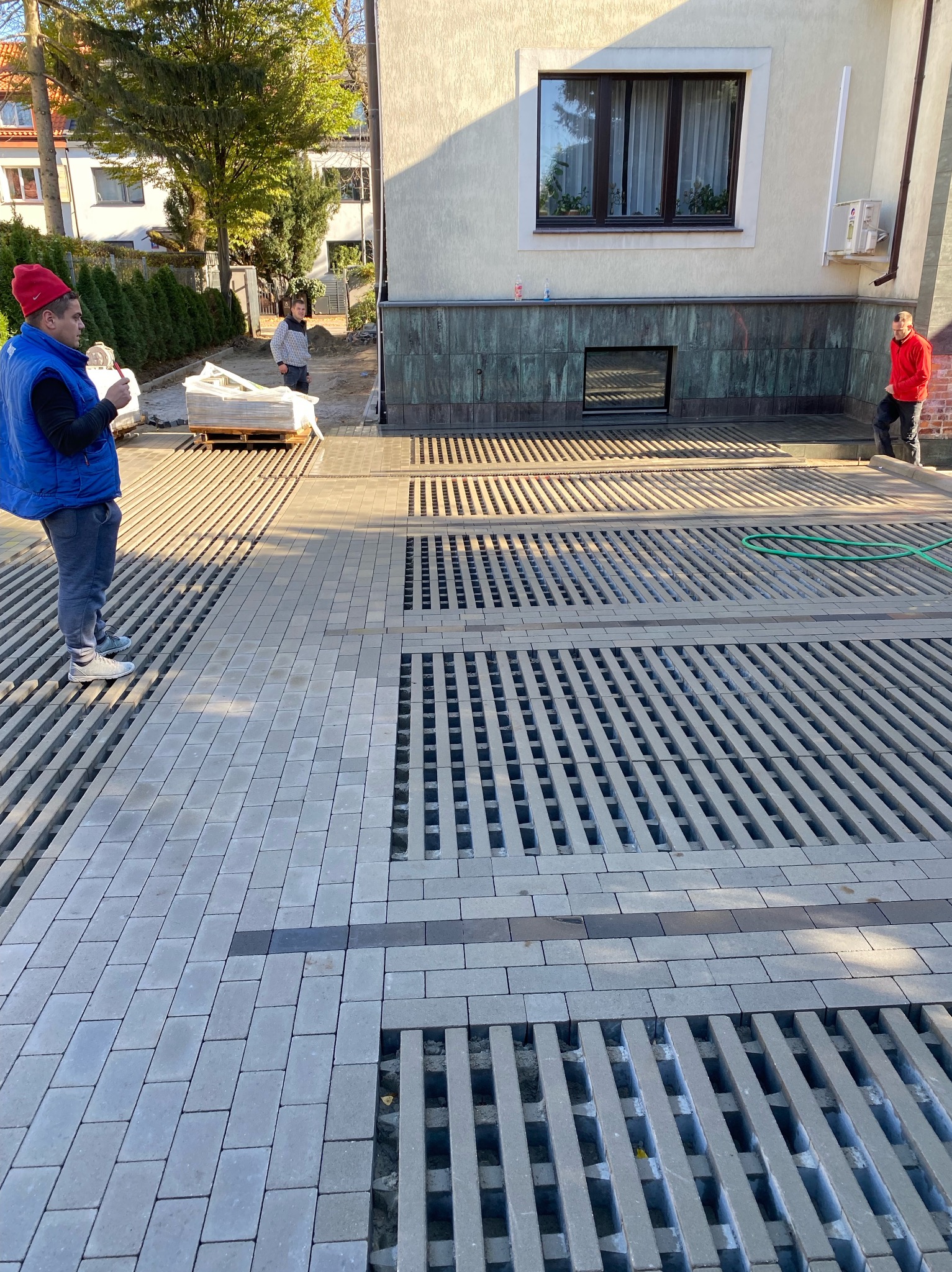 Montaż ażurowej kostki brukowej na podjeździe. Widoczny pracownik w czerwonej czapce i niebieskiej kamizelce. Częściowo ułożona kostka, paleta z kostką w tle.