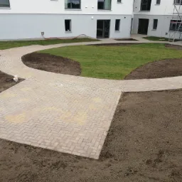 Ukończona ścieżka z kostki brukowej w kształcie łuku, otaczająca trawnik przed budynkiem mieszkalnym w trakcie budowy, widoczne rusztowania.