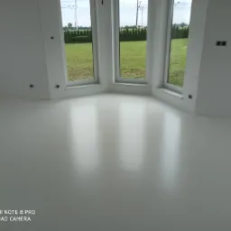 Jasna, jednolita podłoga w nowym pomieszczeniu z trzema oknami narożnymi, przez które widać zielony trawnik i żywopłot.