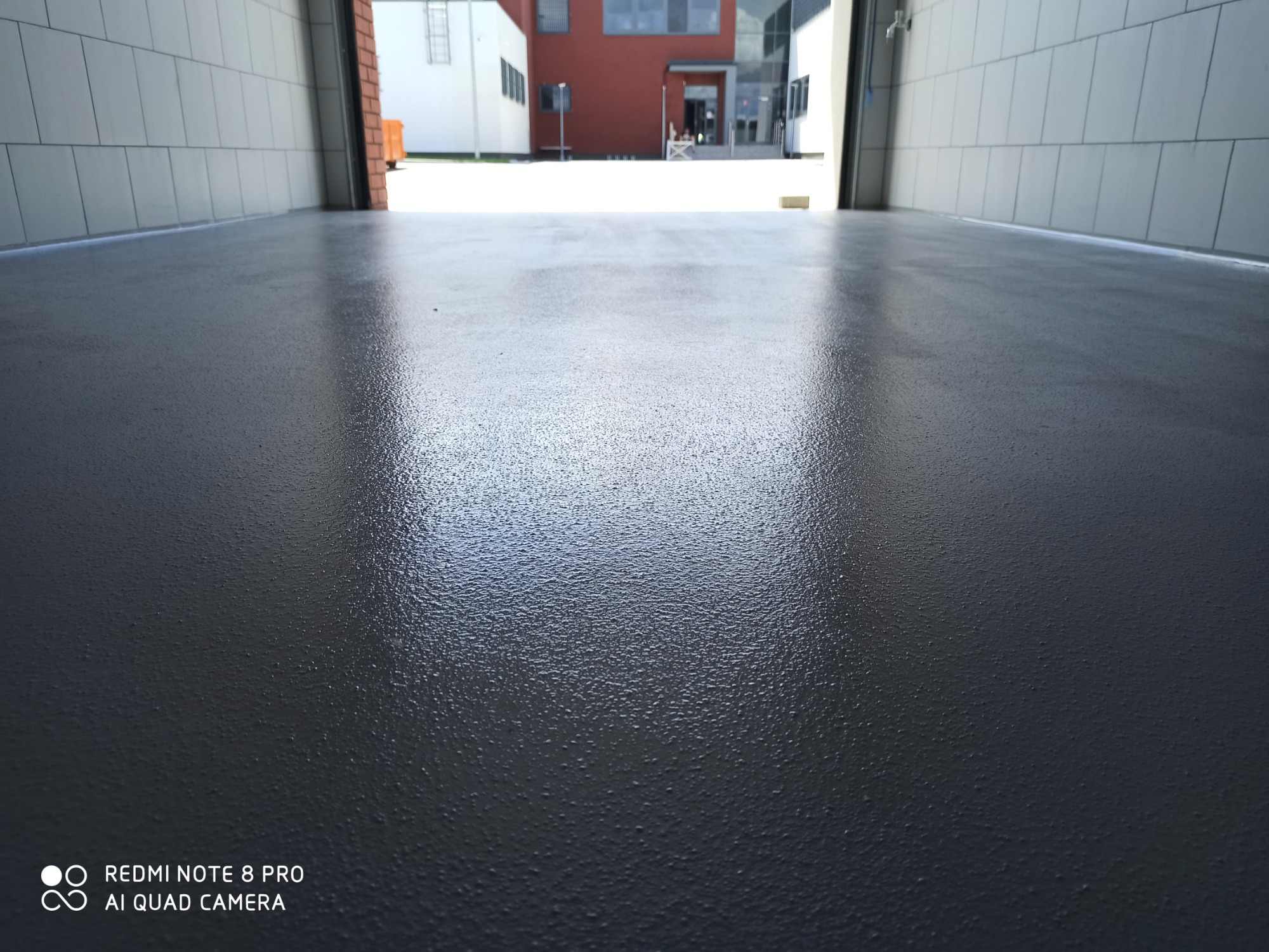 Błyszcząca, szara posadzka żywiczna w garażu lub hali, widok z poziomu podłogi w kierunku otwartej bramy, budynek w tle.
