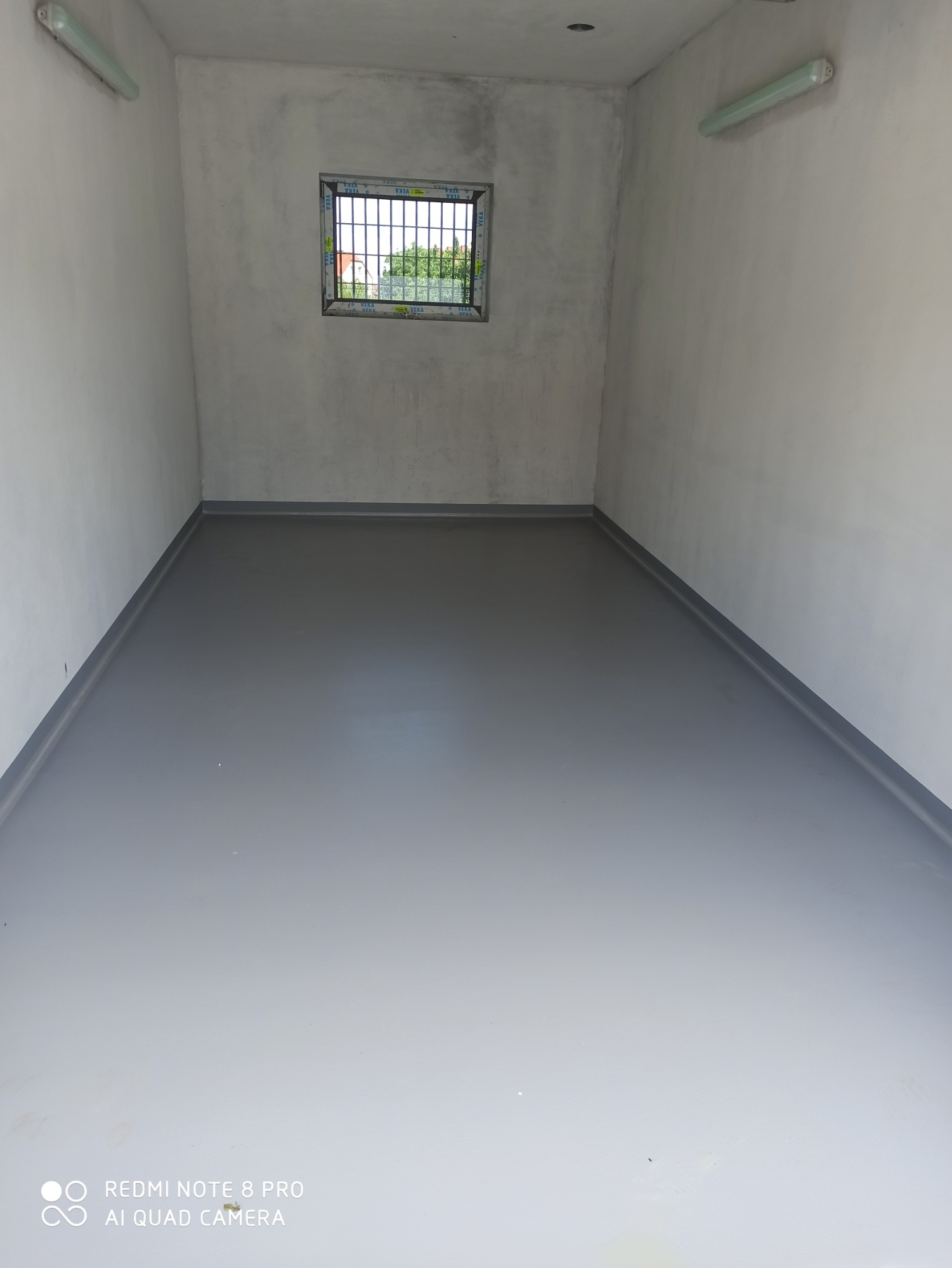Szara, gładka posadzka w pustym pomieszczeniu z oknem zabezpieczonym kratą, widoczne ściany w odcieniach szarości i białe lampy sufitowe.