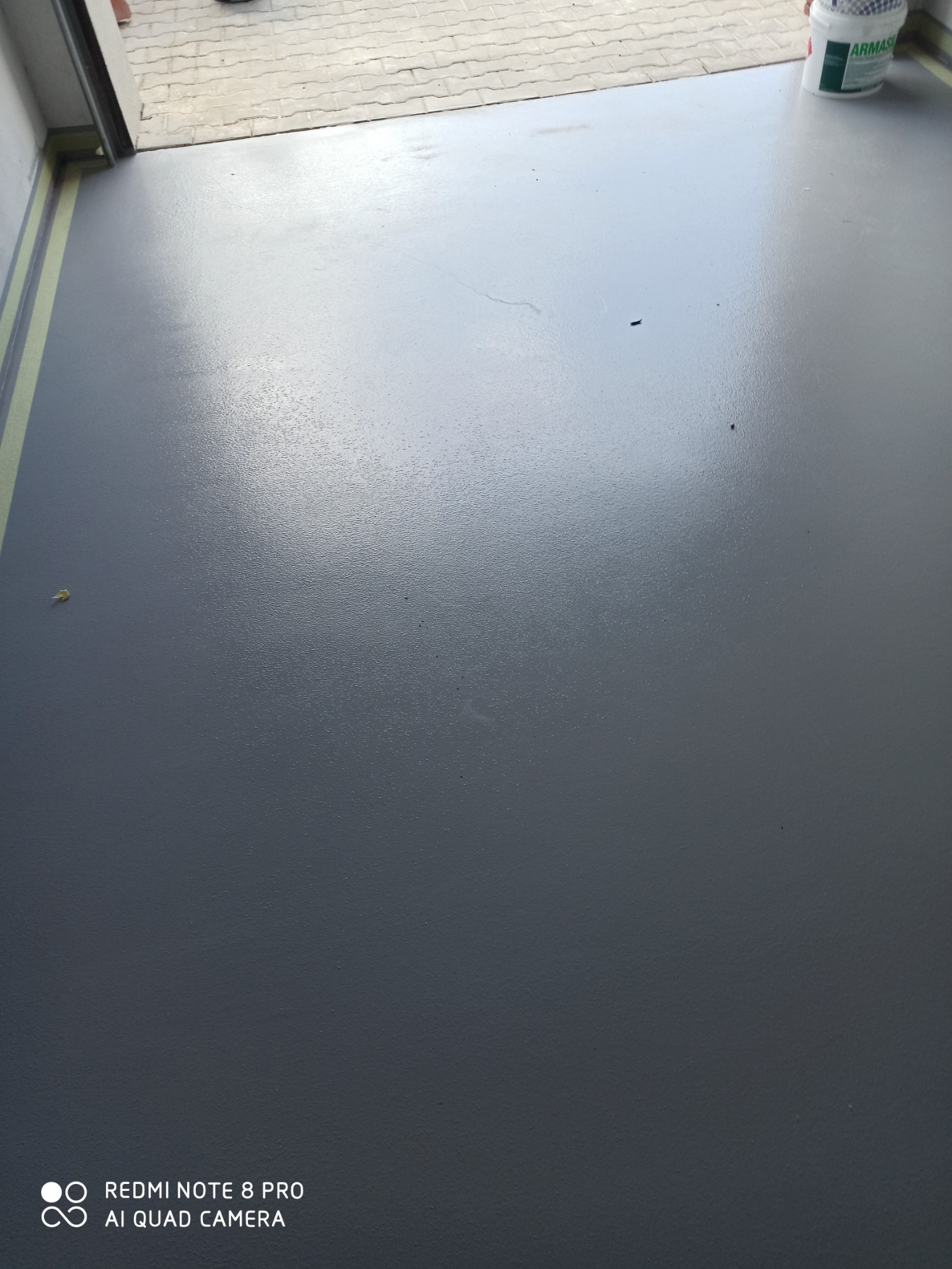 Świeżo wylana, gładka, szara powierzchnia w garażu, z widocznym wiadrem farby w rogu i taśmą maskującą przy ścianie.