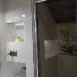 Łazienka po renowacji z beżowymi płytkami, podświetlanymi wnękami, kabiną prysznicową z chromowanymi profilami i dekoracyjnym motywem żaglowca na ścianie.