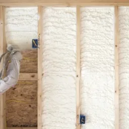 Pracownik w kombinezonie ochronnym aplikuje pianę izolacyjną na ścianę poddasza z drewnianą konstrukcją, widoczne kable elektryczne i puszki instalacyjne.