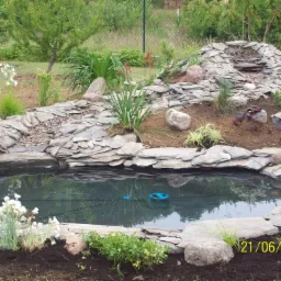 Oczko wodne z kaskadą wyłożoną płaskimi kamieniami, otoczone roślinami ozdobnymi i kamieniami, z widocznym niebieskim pływakiem w wodzie.