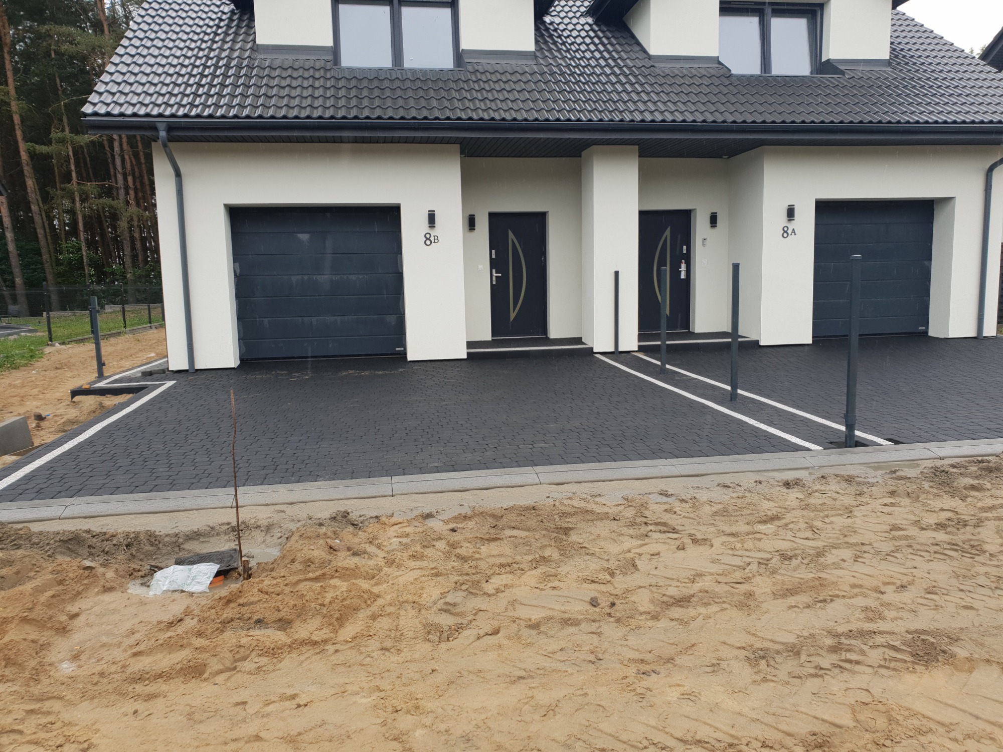 Nowoczesny bliźniak z ciemną kostką brukową, widoczne przygotowania do dalszych prac ziemnych, z fragmentem wykopu i wystającą rurą.