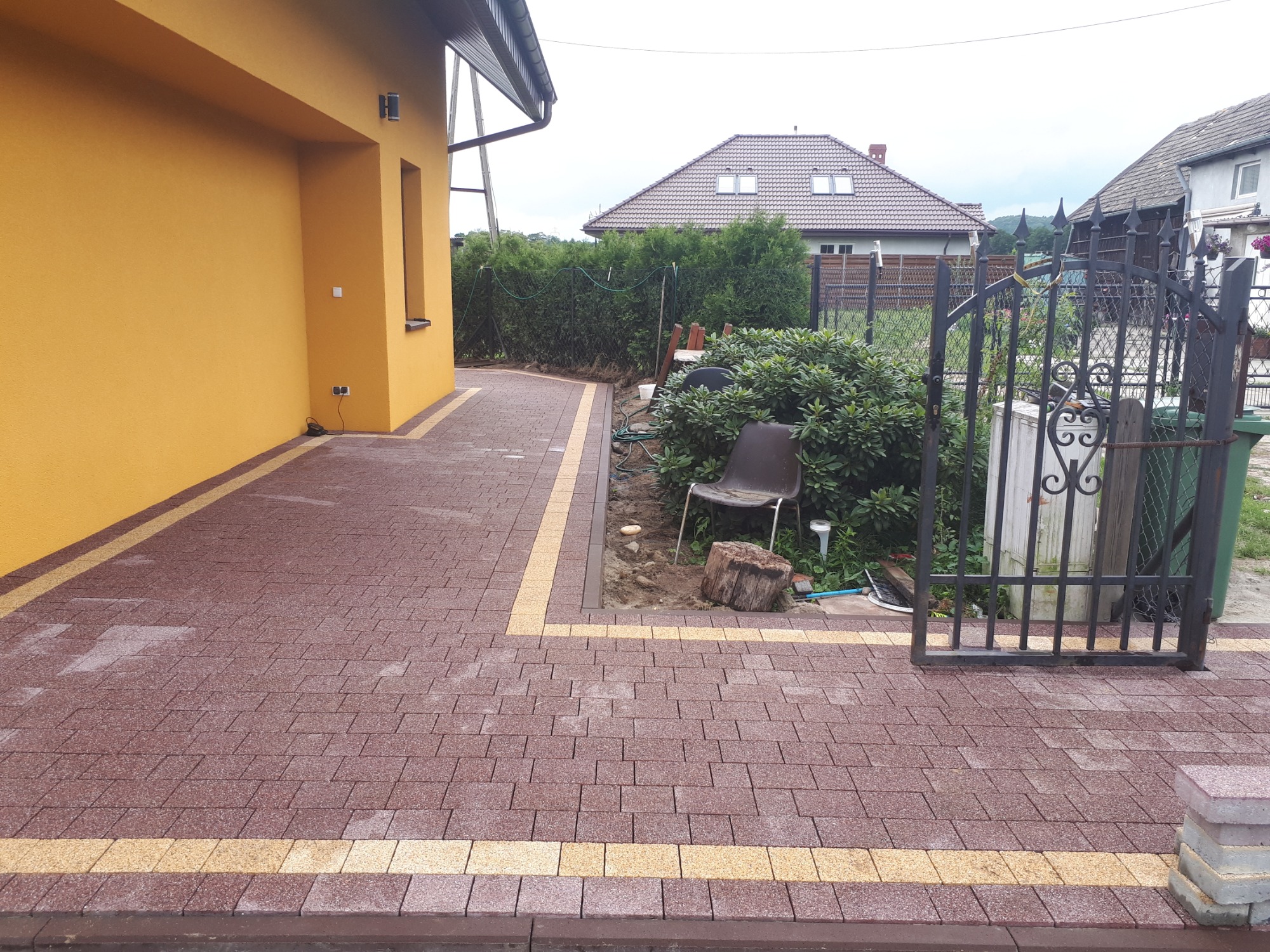 Nowo ułożona, brązowo-żółta kostka brukowa przed żółtym budynkiem, z widocznym fragmentem ogrodu z metalową bramką i krzesłem w tle.
