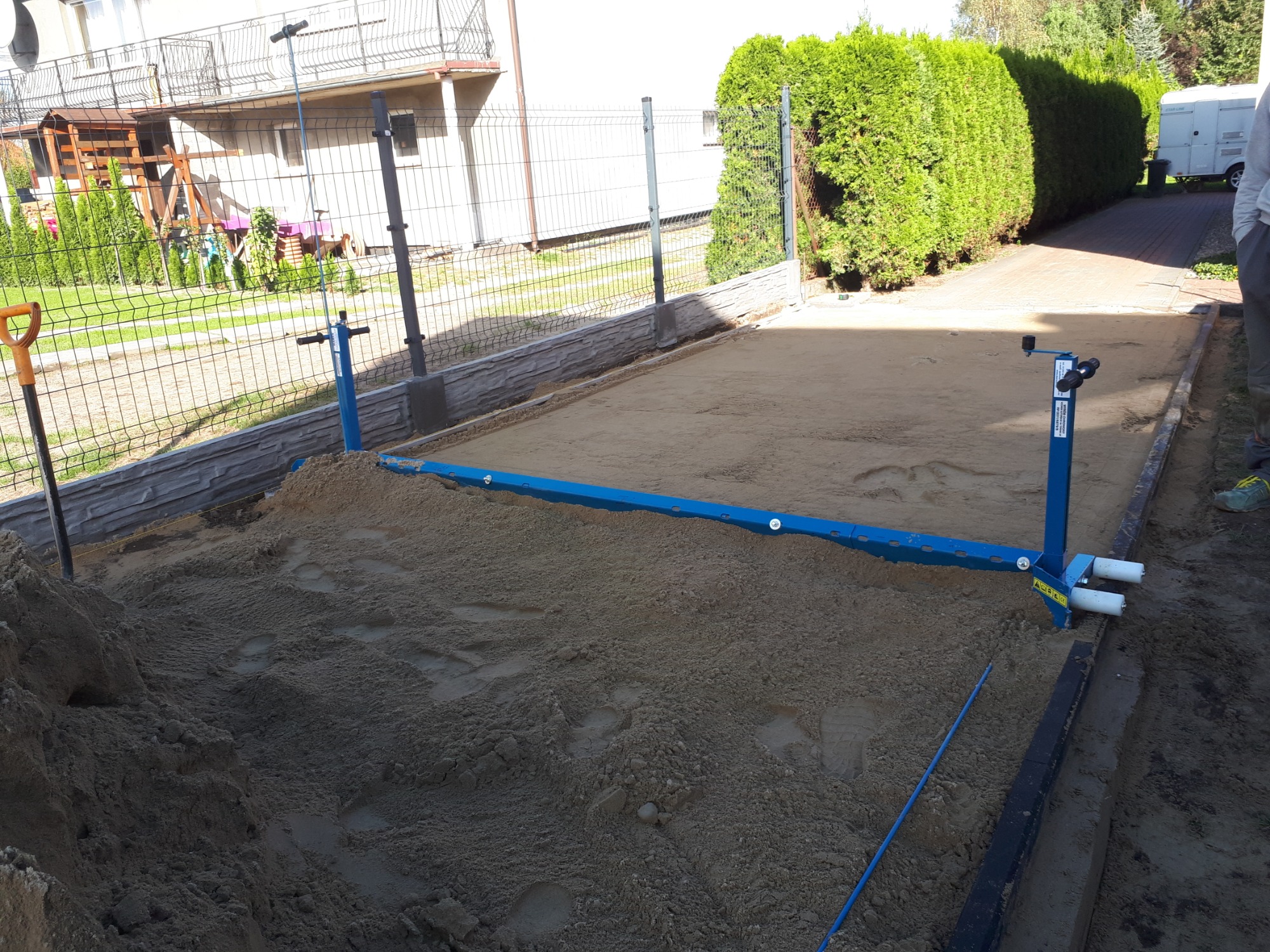 Przygotowanie podłoża z piasku pod kostkę brukową, widoczne narzędzia do poziomowania, obrzeża betonowe i ogrodzenie panelowe.