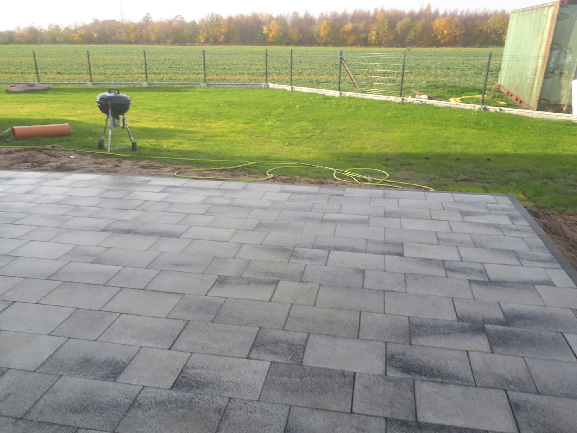 Nowo ułożona, szara kostka brukowa na posesji z widokiem na trawnik, ogrodzenie i pole w tle. Na trawniku widoczny grill, rura PCV i zwinięty wąż ogrodowy.