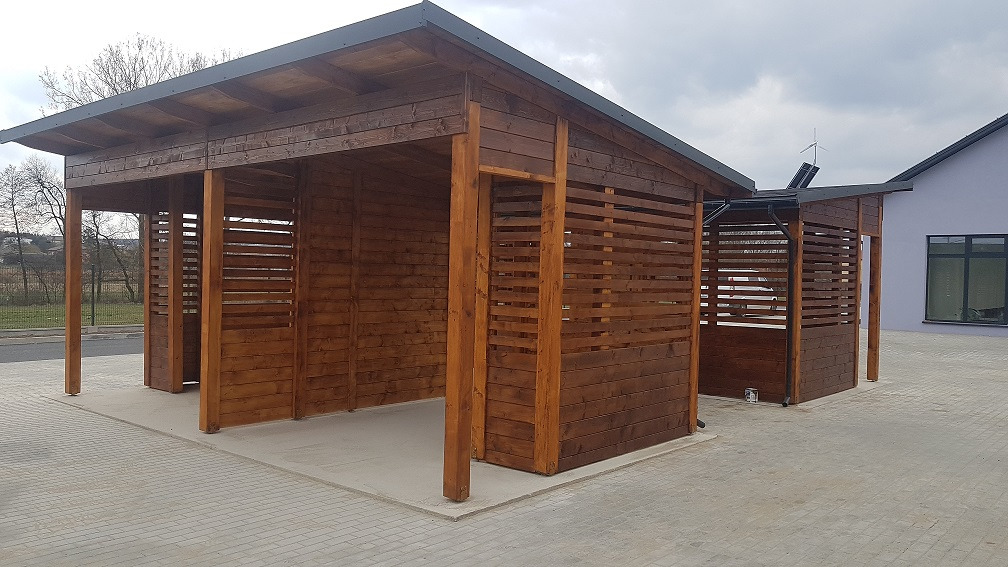 Dwie drewniane altany z poziomymi deskami, połączone z budynkiem z szarą elewacją, widok z zewnątrz na brukowany plac.