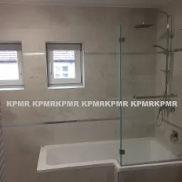 Wykończona łazienka z wanną obudowaną płytkami, szklanym parawanem nawannowym, deszczownicą i dwoma oknami.