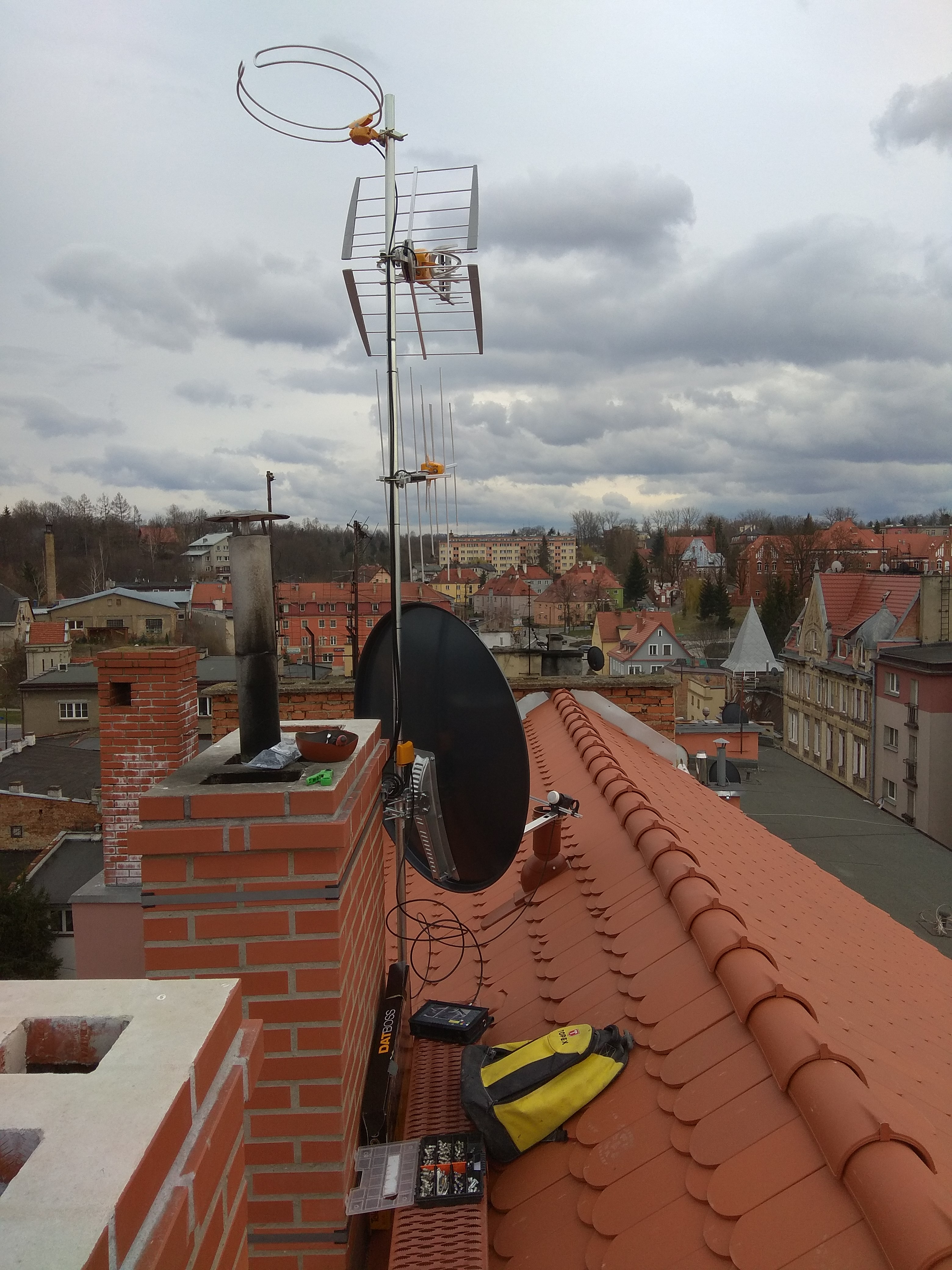 Antena telewizyjna i satelitarna zamontowana na dachu budynku z czerwonej dachówki, widok z góry na miasto pod pochmurnym niebem, narzędzia i torba monterska leżą obok komina.
