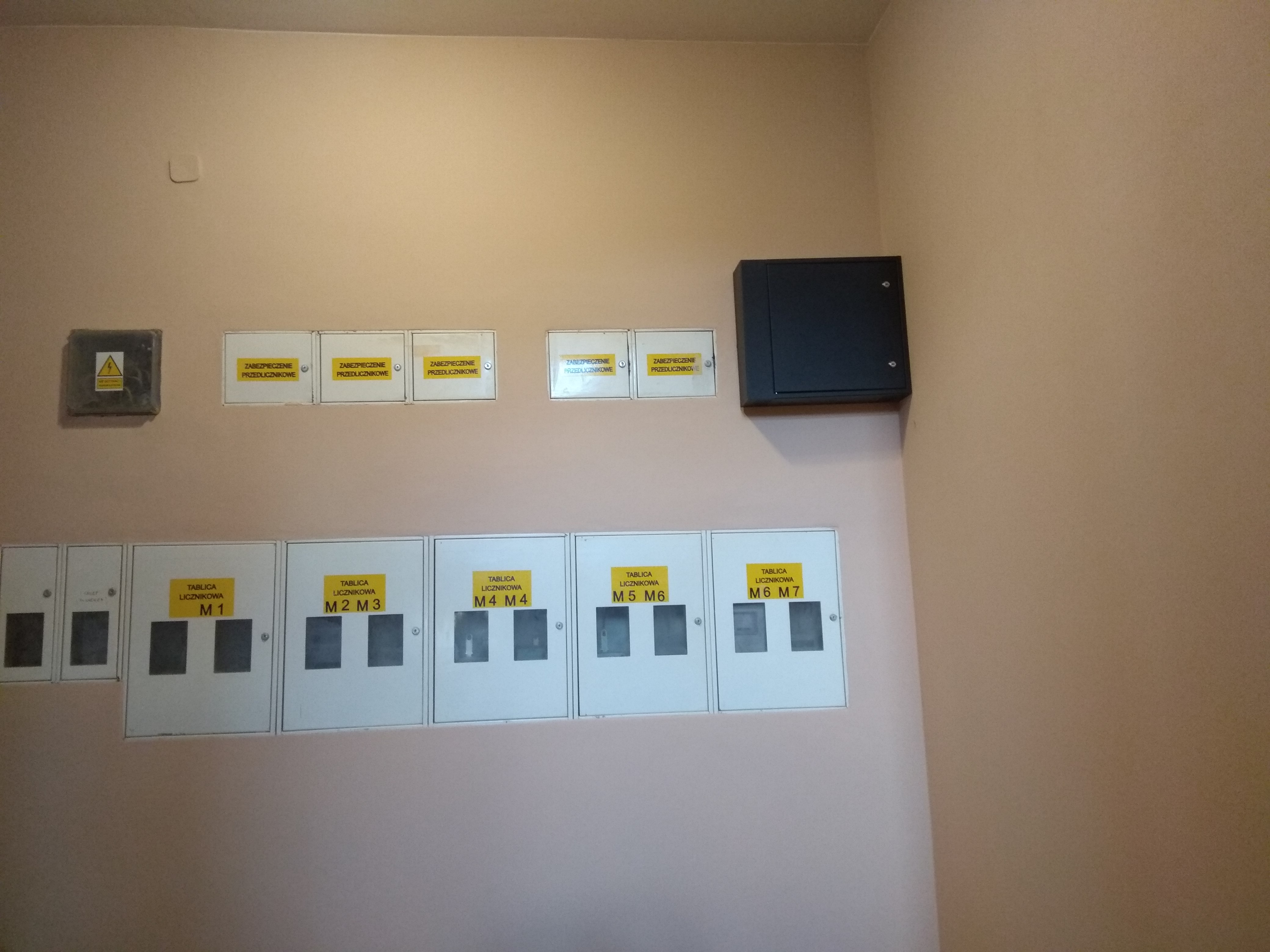 Tablica rozdzielcza z kilkoma bezpiecznikami i tabliczkami licznikowymi M1 do M7 na beżowej ścianie, dodatkowo czarna skrzynka rozdzielcza z prawej strony.