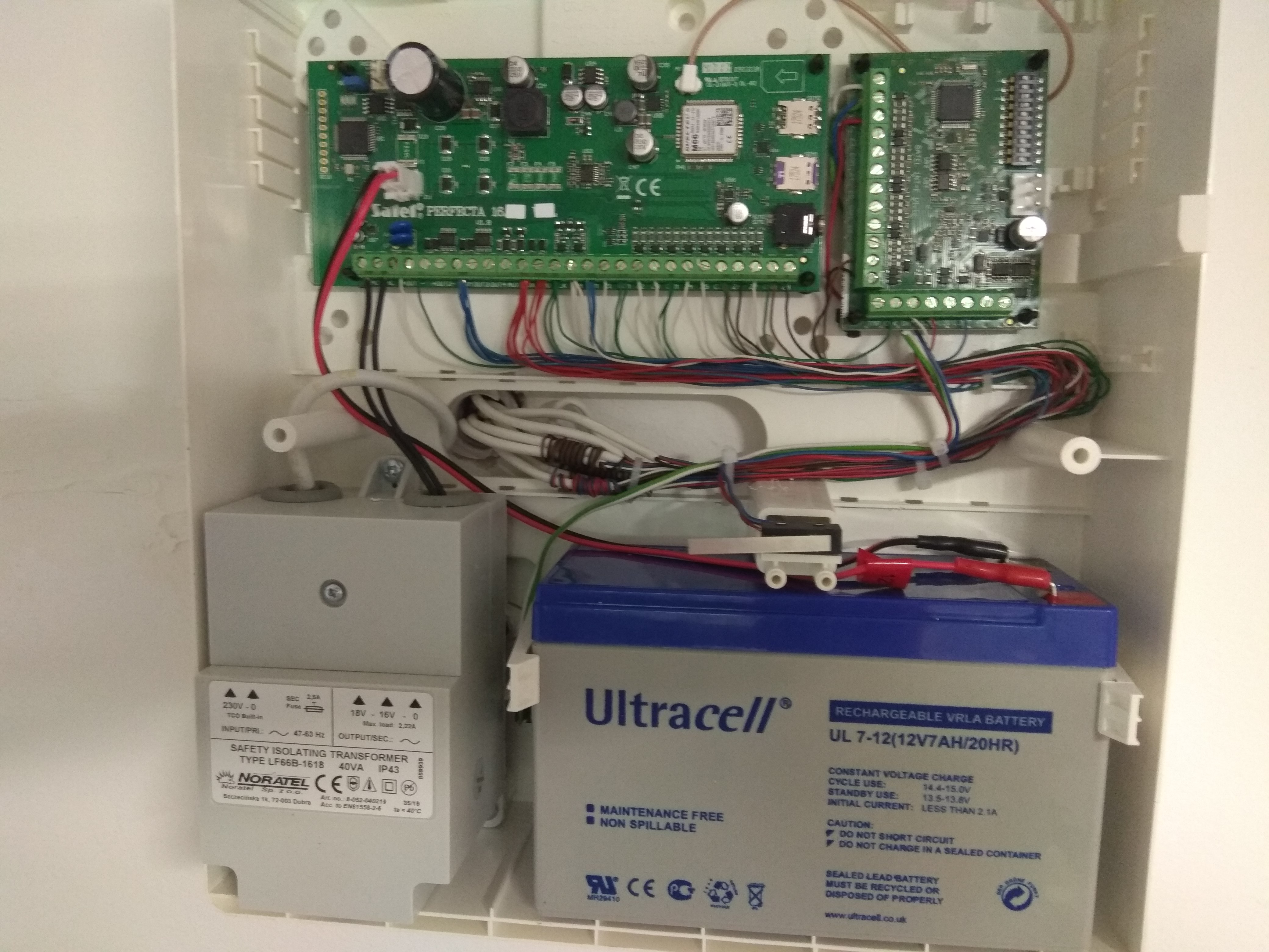 Otwarta skrzynka z elektroniką systemu alarmowego: dwie zielone płytki drukowane, akumulator Ultracell i transformator Noratel, połączone przewodami w kolorach czerwonym, niebieskim i czarnym.