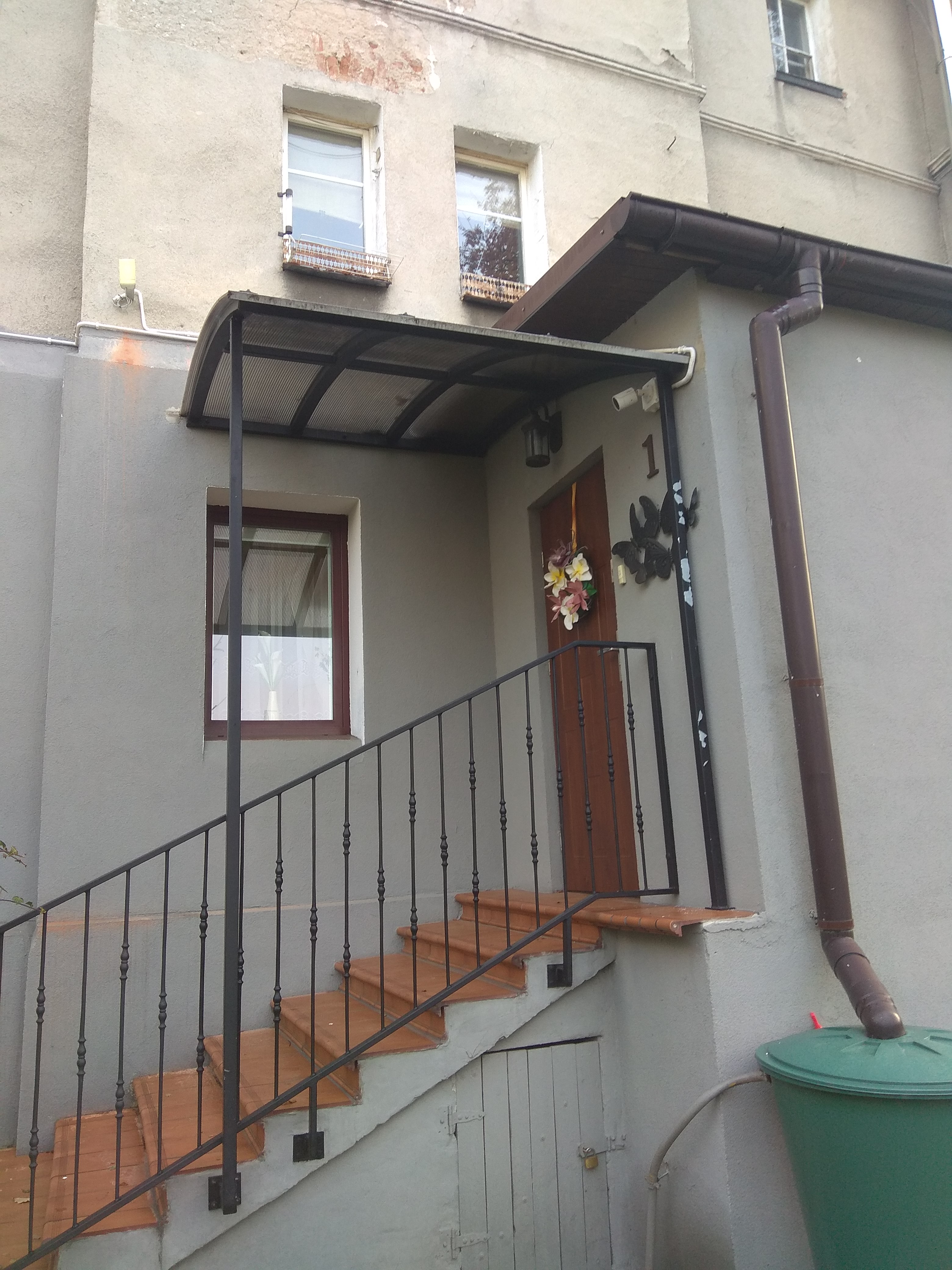 Wejście do budynku z brązowymi schodami, czarną metalową balustradą i zadaszeniem, ozdobione kwiatowym wieńcem na drzwiach i metalowymi motylami obok numeru domu.