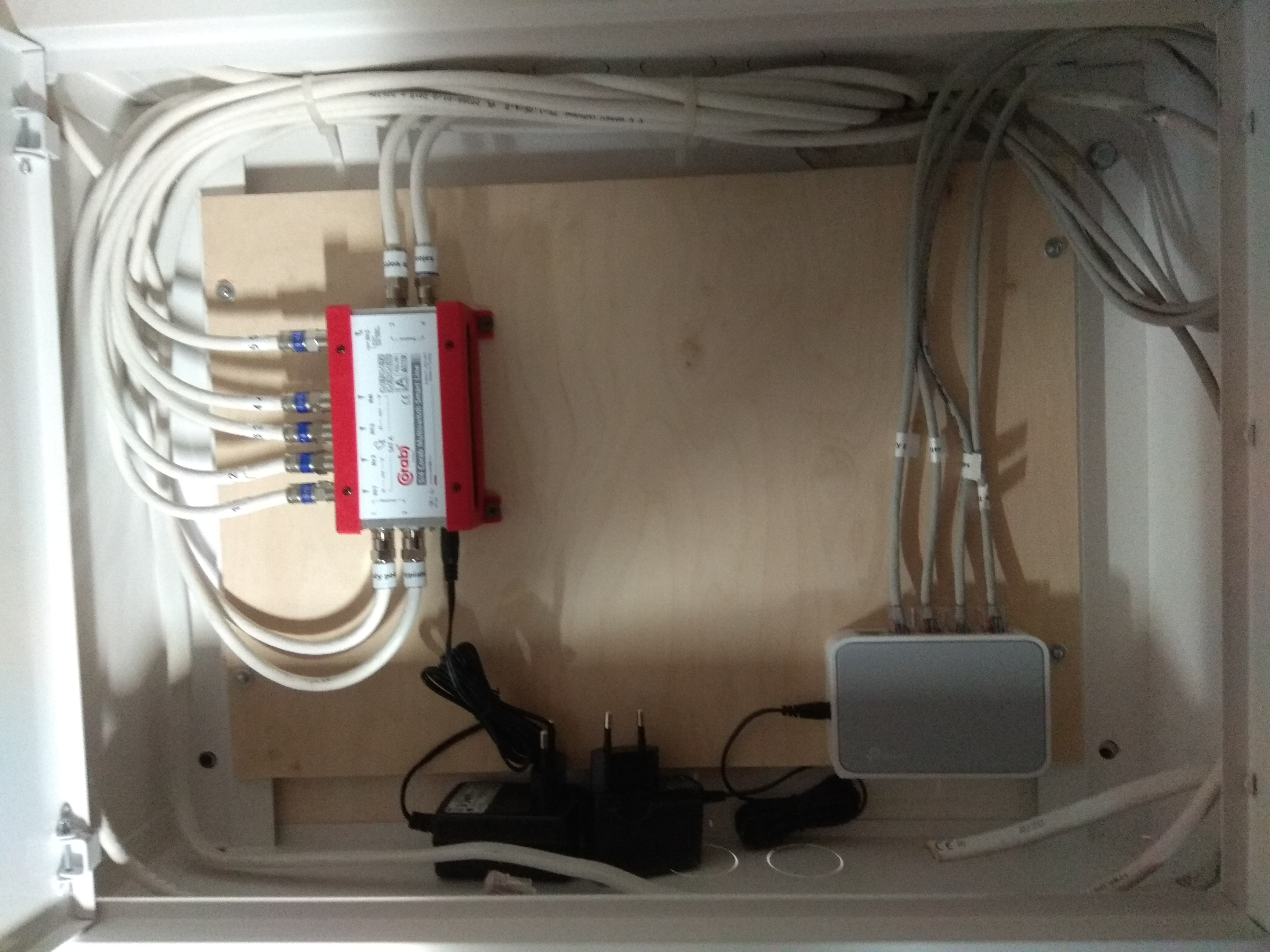 Rozdzielacz sygnału TV z podłączonymi kablami koncentrycznymi i zasilaczem wewnątrz szafki, obok router sieciowy z kablami.