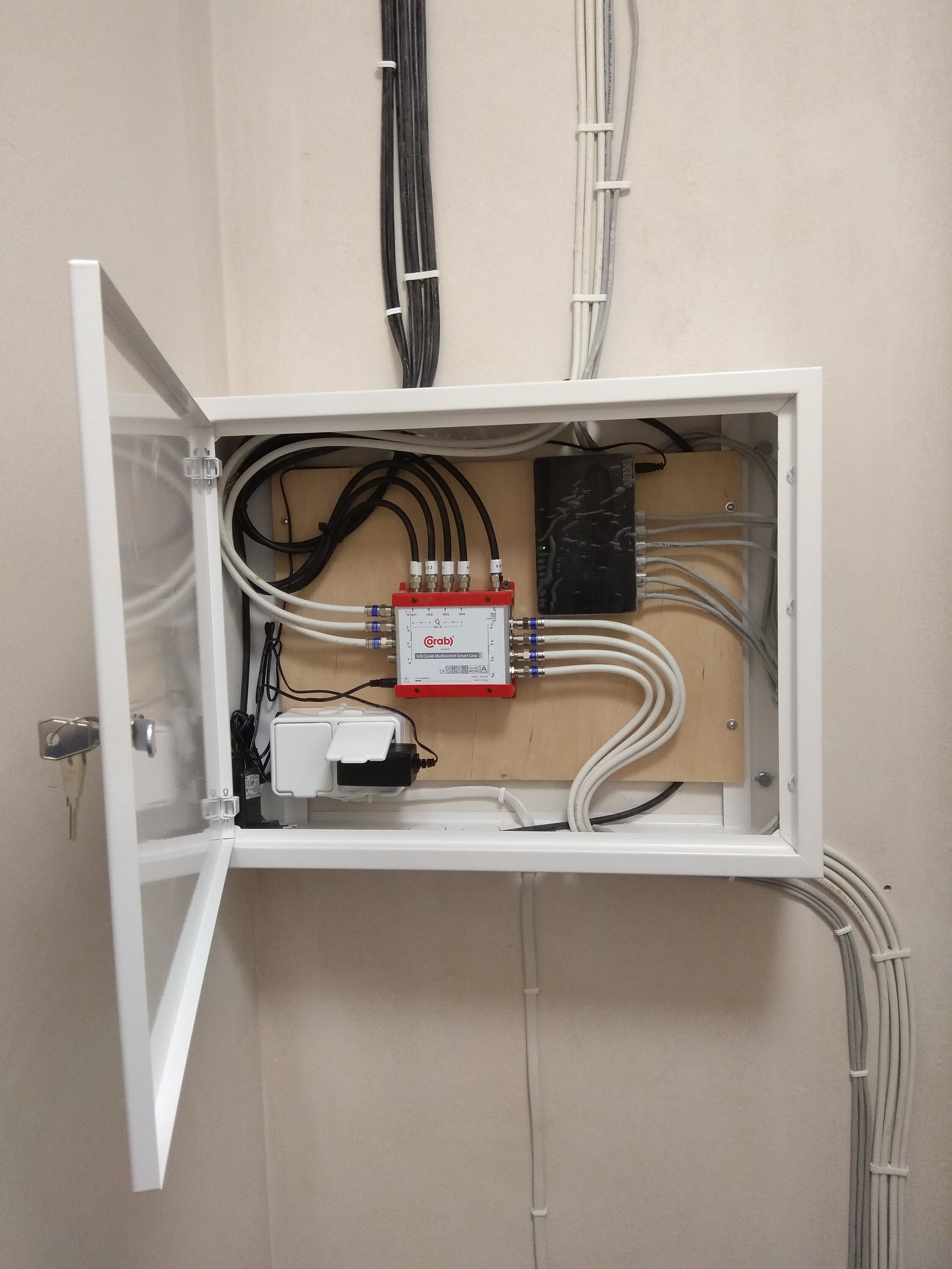 Otwarta skrzynka z rozdzielaczem sygnału telewizyjnego i routerem, widoczne kable przymocowane do ściany, klucz w zamku drzwi skrzynki.