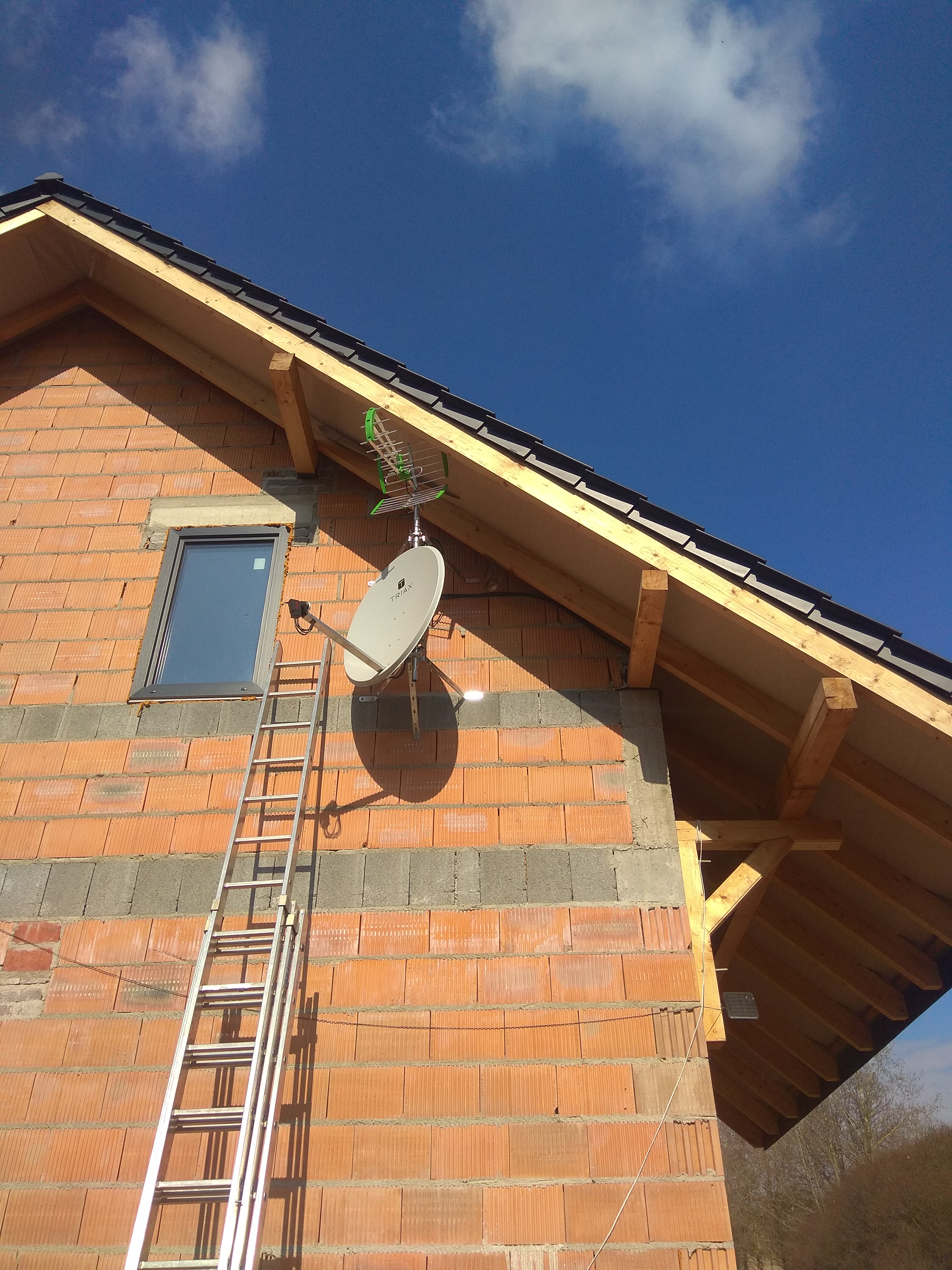Antena satelitarna Triax i antena naziemna zamontowane na bocznej ścianie domu w trakcie budowy, aluminiowa drabina oparta o ścianę, błękitne niebo w tle.