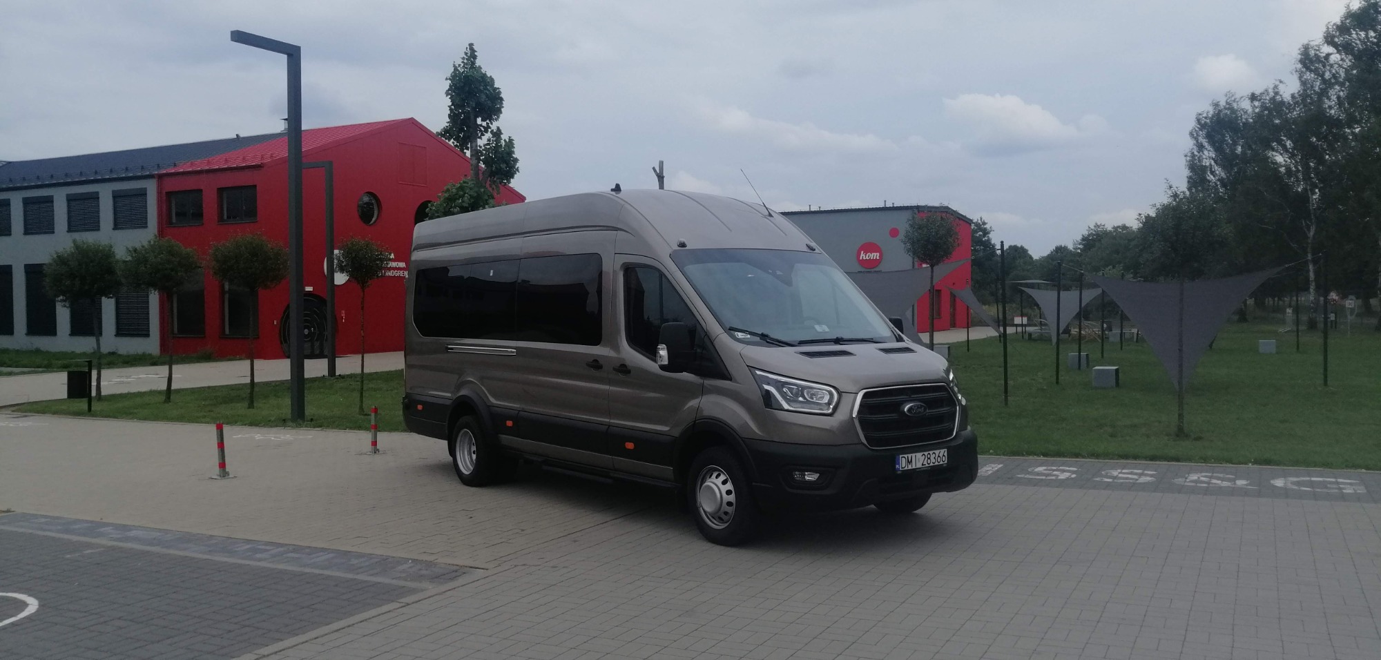 Szary bus Ford Transit na tle nowoczesnego budynku z czerwonymi elementami, widoczne tablice rejestracyjne.