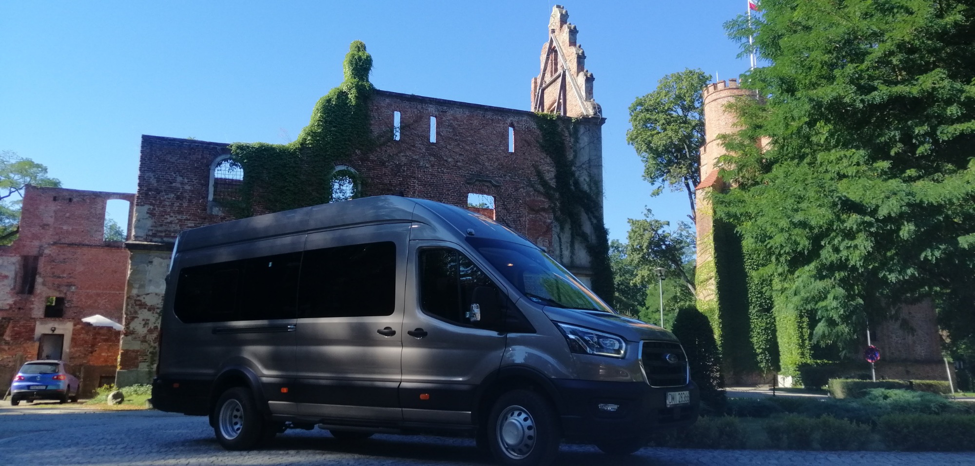 Szary bus transportowy na tle ruin zamku obrośniętego bluszczem w Bukowicach.