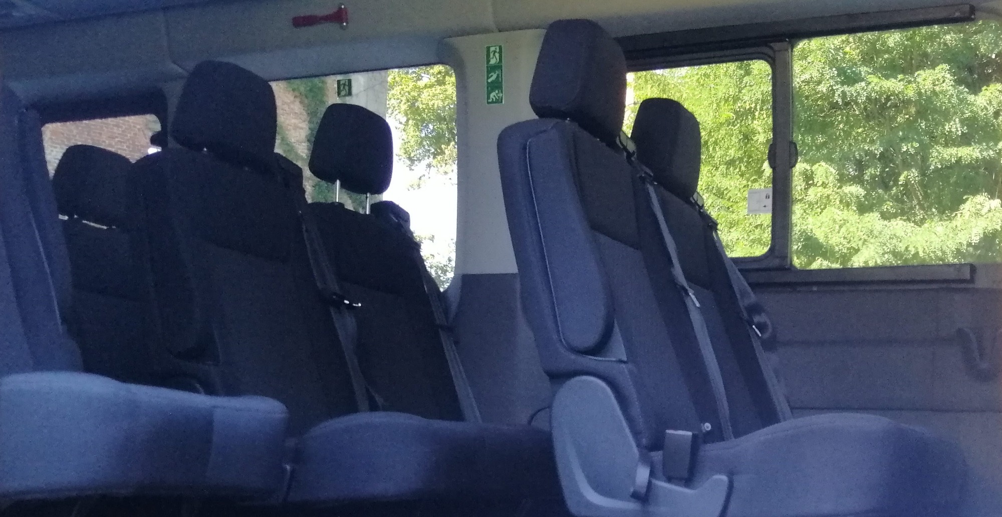 Wnętrze busa z rzędami ciemnych foteli pasażerskich, widok przez okno na zieleń drzew.