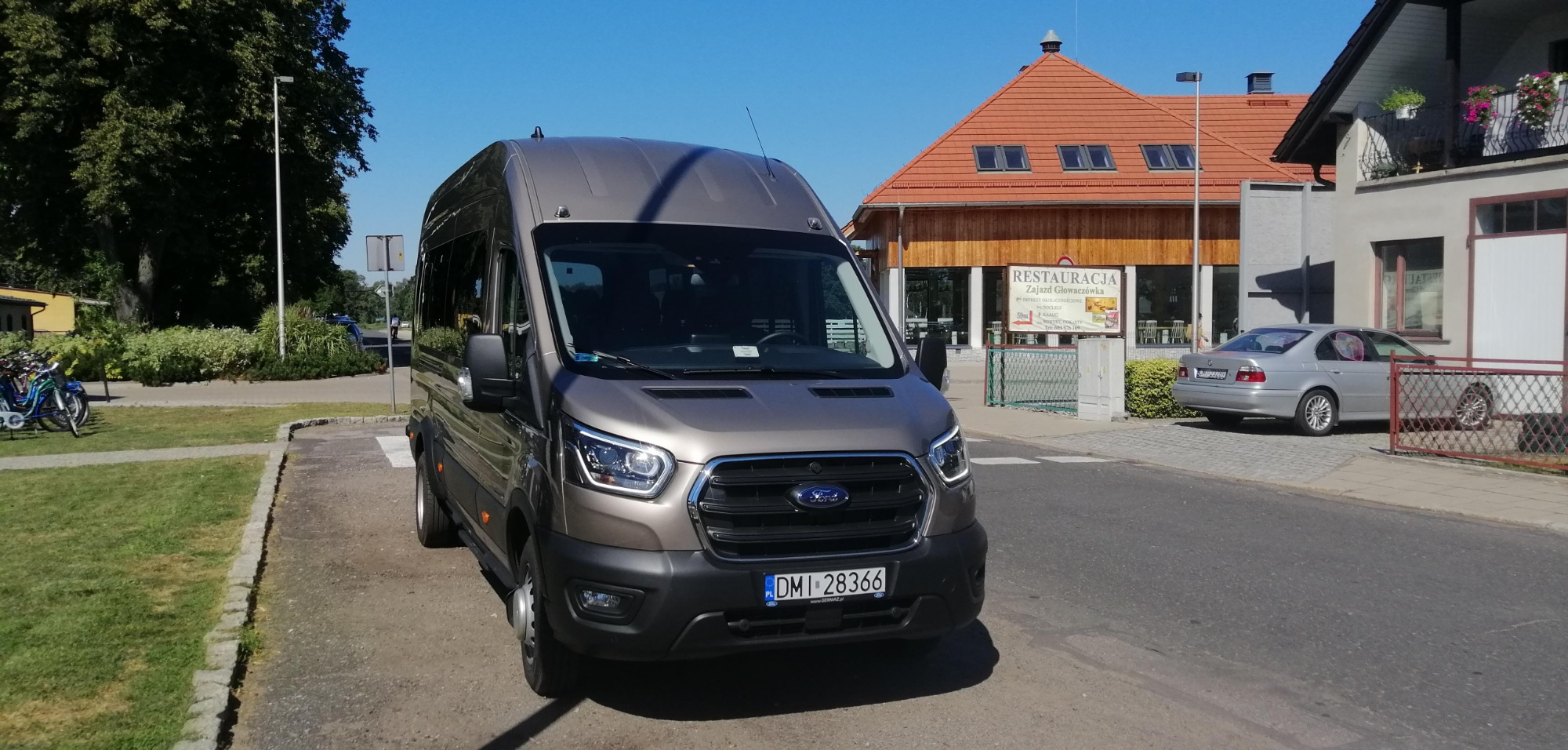 Szary Ford Transit na przejściu dla pieszych przed Zajazdem Głowaczówka w słoneczny dzień.