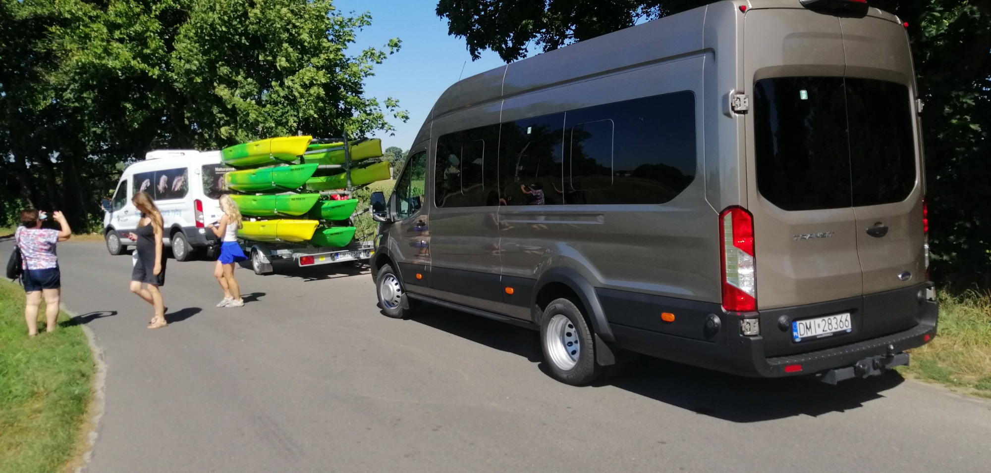 Szary bus z przyczepą wypełnioną zielono-żółtymi kajakami na drodze, w tle biały bus z grafiką zwierząt i spacerujące osoby.