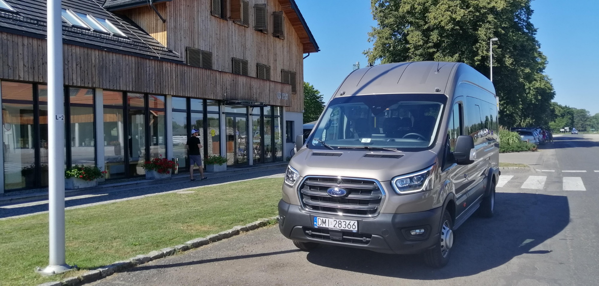 Szary bus Ford Transit z numerem rejestracyjnym DM 28366 zaparkowany przed budynkiem z drewnianą elewacją i szklanymi witrynami.