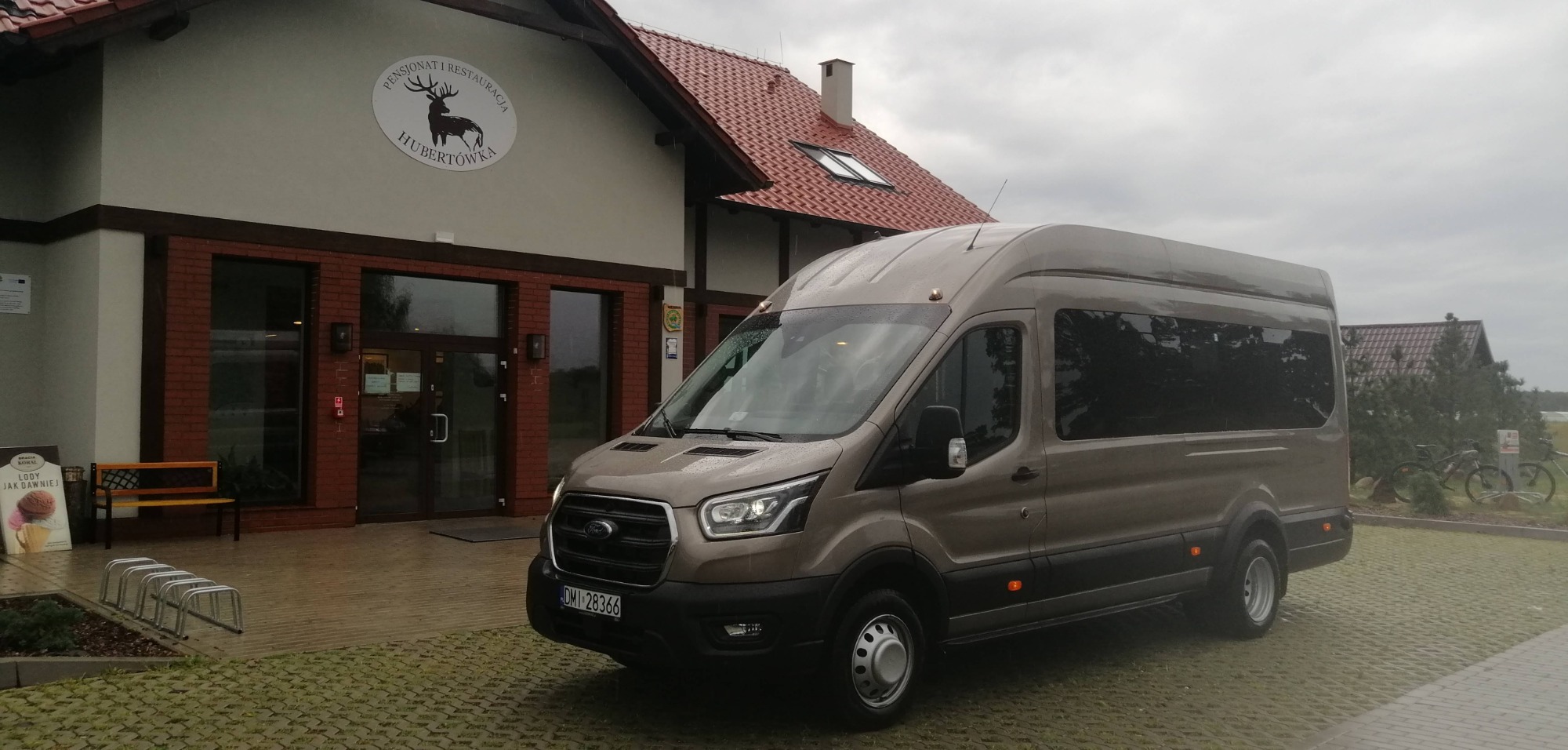 Brązowy bus Ford Transit z przyciemnianymi szybami zaparkowany przed budynkiem Pensjonatu i Restauracji Hubertówka w pochmurny dzień.