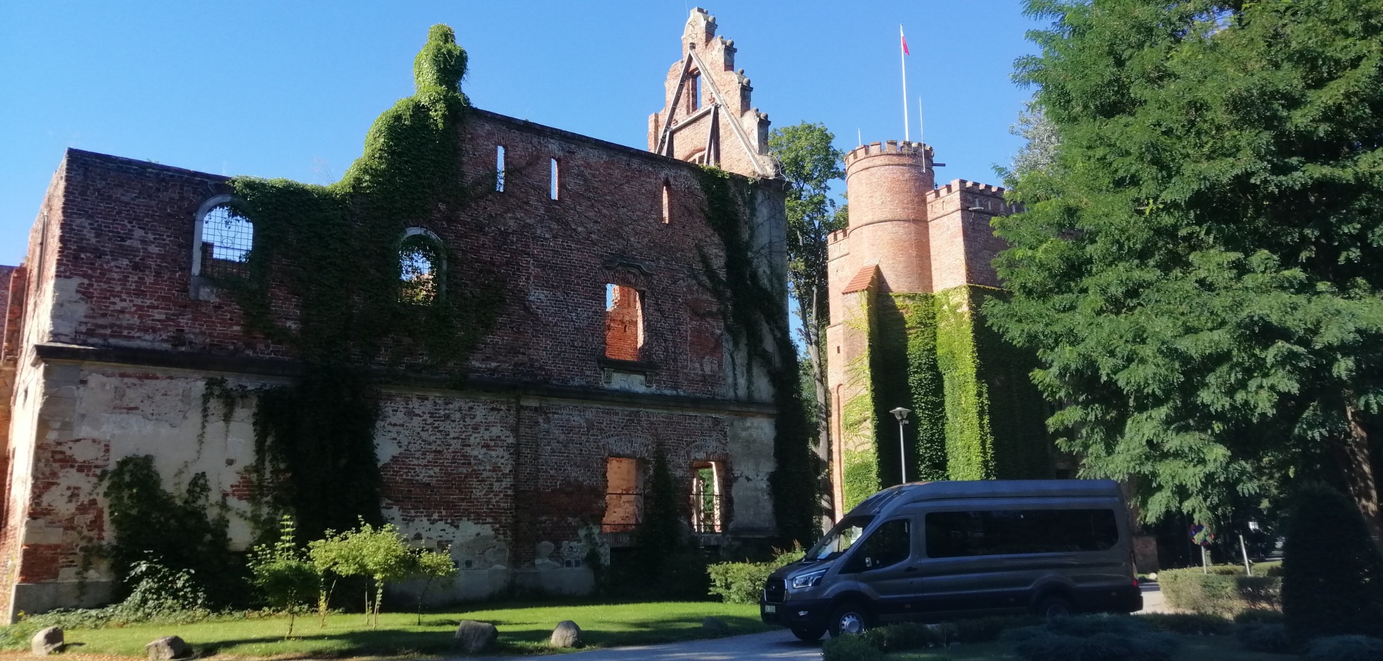 Szary bus na tle ceglanych ruin zamku obrośniętych bluszczem i wieży z flagą na szczycie w słoneczny dzień.