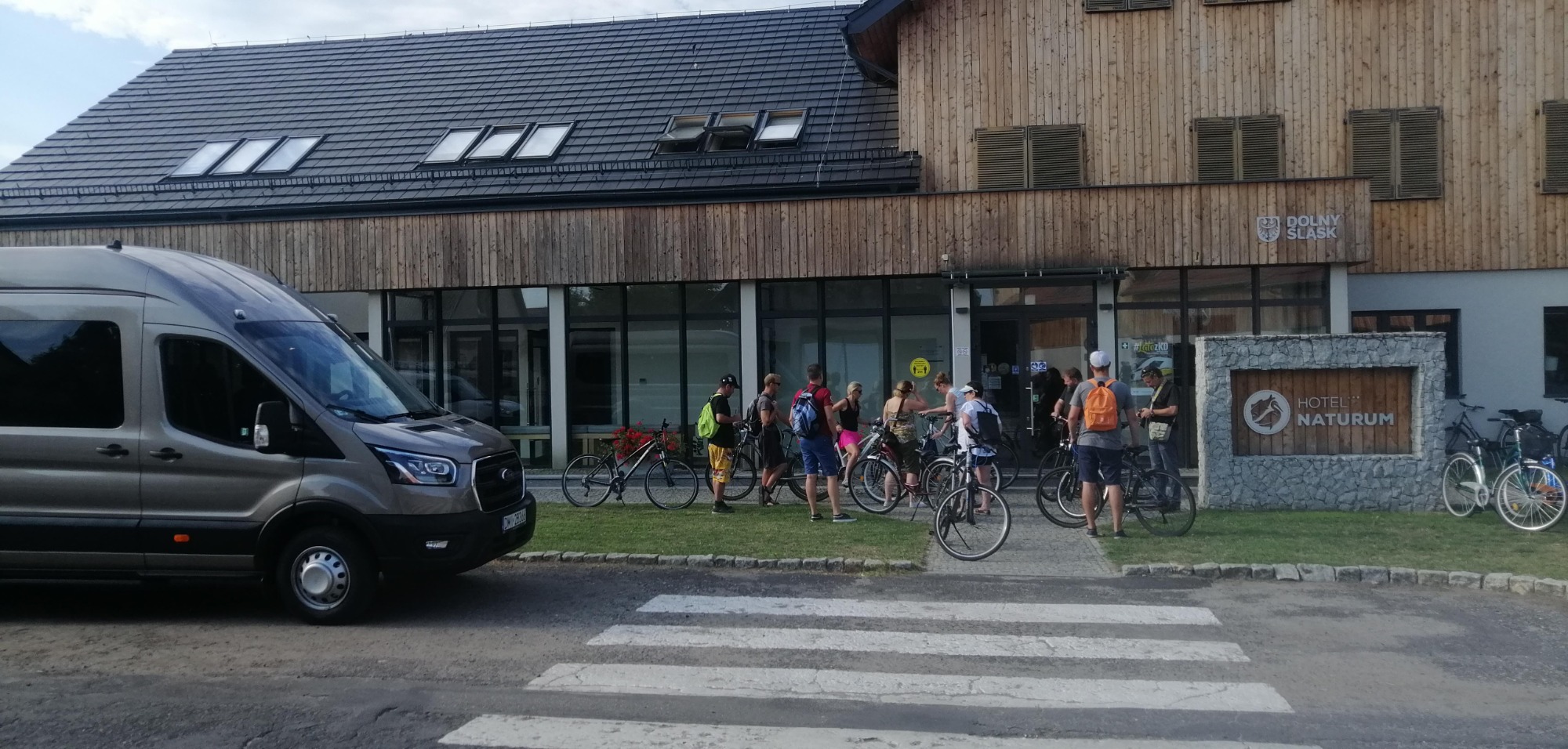 Szary bus transportowy zaparkowany przed budynkiem Hotelu Naturum, grupa osób z rowerami w tle, przejście dla pieszych na pierwszym planie.
