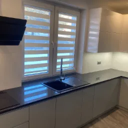 Nowoczesna kuchnia z ciemnym, nakrapianym blatem, czarnym zlewem podwieszanym i płytą indukcyjną, jasne lakierowane szafki i okno z roletami dzień-noc.