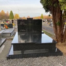 Czarny, granitowy pomnik z wyrytym imieniem i nazwiskiem Julian Ordyna oraz datami urodzin i śmierci, otoczony szarym żwirem i wysokim drzewem iglastym na cmentarzu.