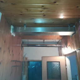 Wnętrze wykończone boazerią z widocznym kanałem wentylacyjnym pod sufitem, rurą biegnącą wzdłuż ściany oraz kratką wentylacyjną z pianką montażową w rogu pomieszczenia.