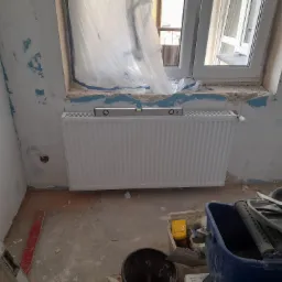 Montaż nowego grzejnika panelowego pod oknem, poziomica na górze, ściany w trakcie remontu, widoczne ślady po starej farbie i przygotowaniu do malowania.
