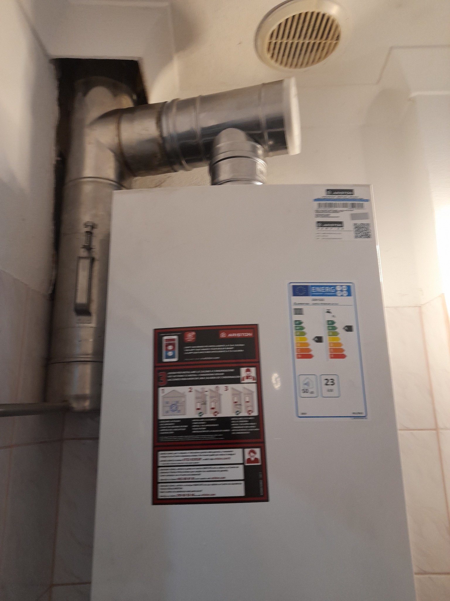 Biały piec gazowy marki Ariston z podłączonymi metalowymi rurami odprowadzającymi spaliny, widoczna etykieta energetyczna i instrukcja obsługi, na ścianie płytki ceramiczne.