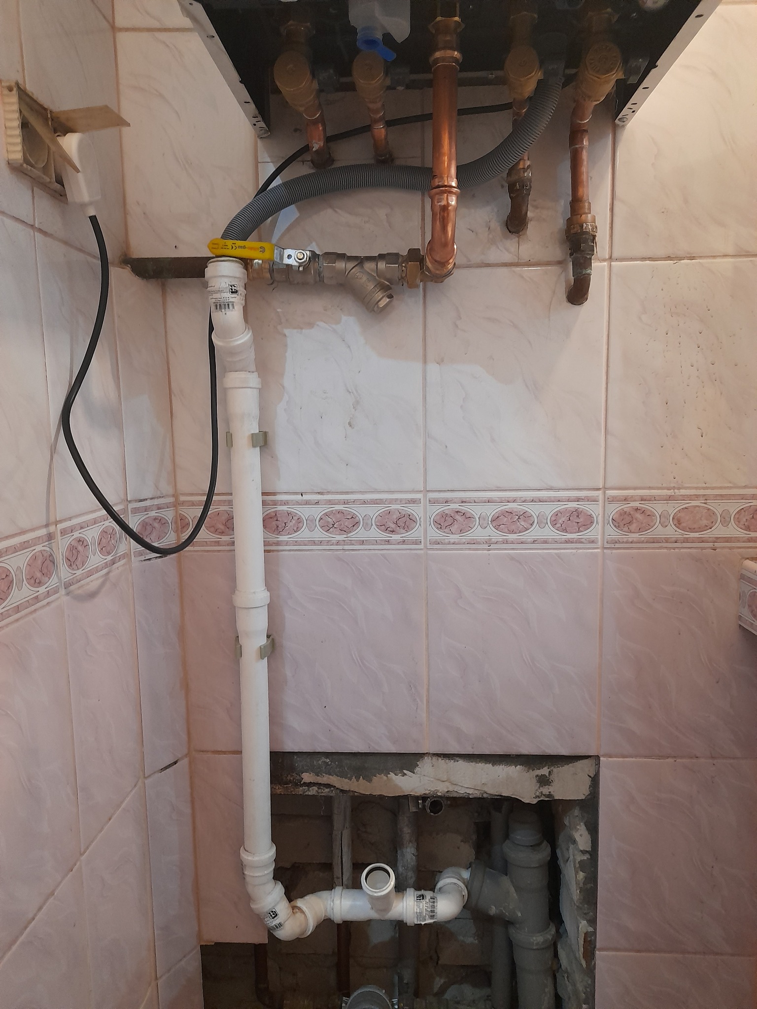 Podłączenie kotła gazowego z widocznymi rurami miedzianymi i plastikowymi, zaworem kulowym z żółtą rączką oraz fragmentem instalacji ukrytym w ścianie z uszkodzonymi płytkami.