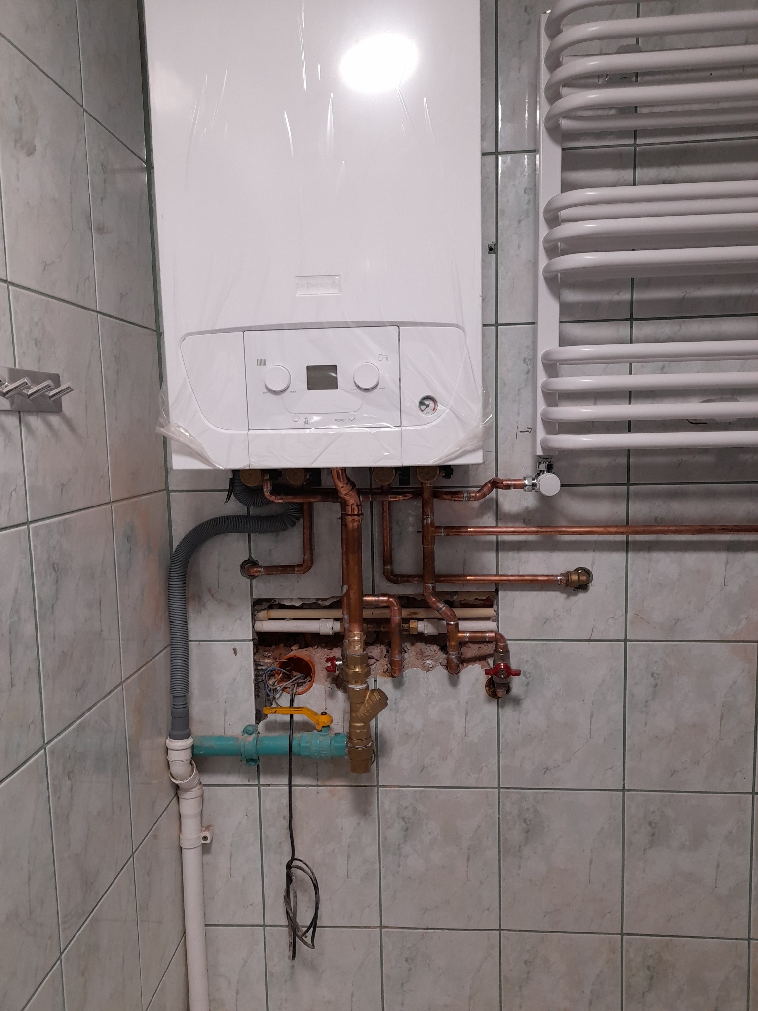 Biały, nowy piec gazowy, jeszcze w folii ochronnej, zamontowany na ścianie z szarych płytek. Widoczne miedziane rury, grzejnik drabinkowy i fragment instalacji wodnej z zaworem.
