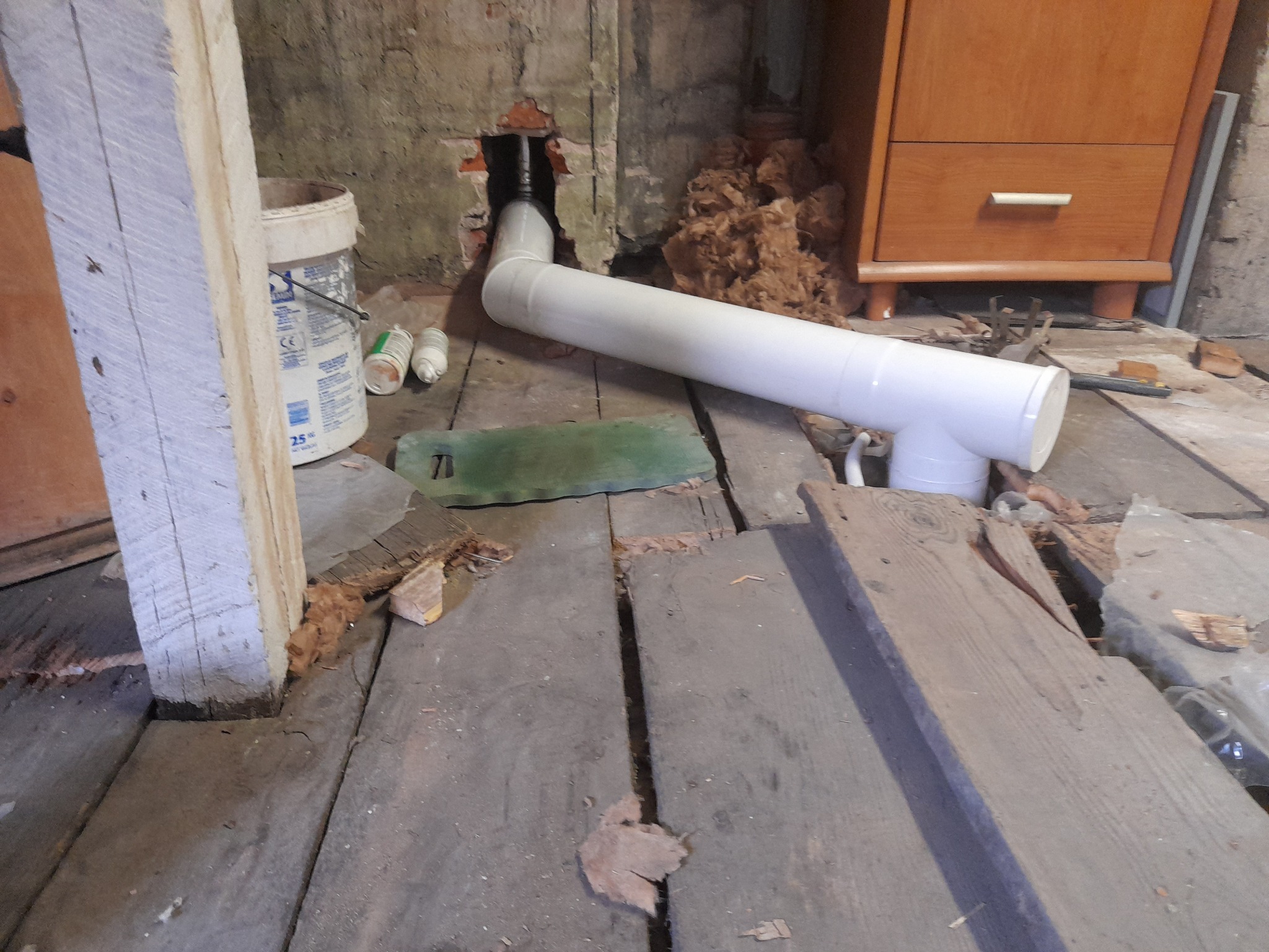 Biała rura PCV wystająca ze ściany z cegły, z elementem T, na tle drewnianej podłogi w trakcie remontu, widoczny fragment komody i puszka farby.