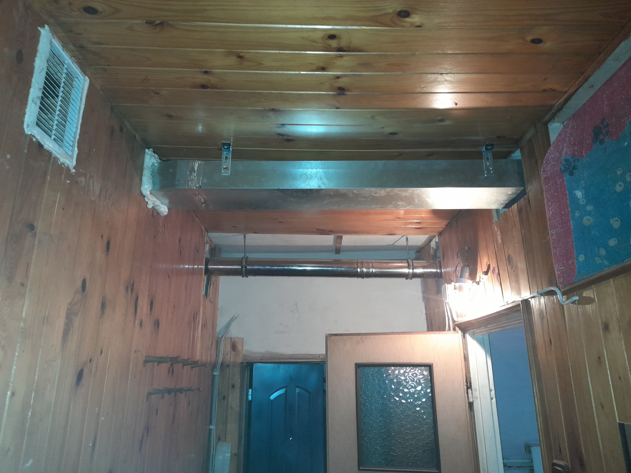 Wnętrze wykończone boazerią z widocznym kanałem wentylacyjnym pod sufitem, rurą biegnącą wzdłuż ściany oraz kratką wentylacyjną z pianką montażową w rogu pomieszczenia.