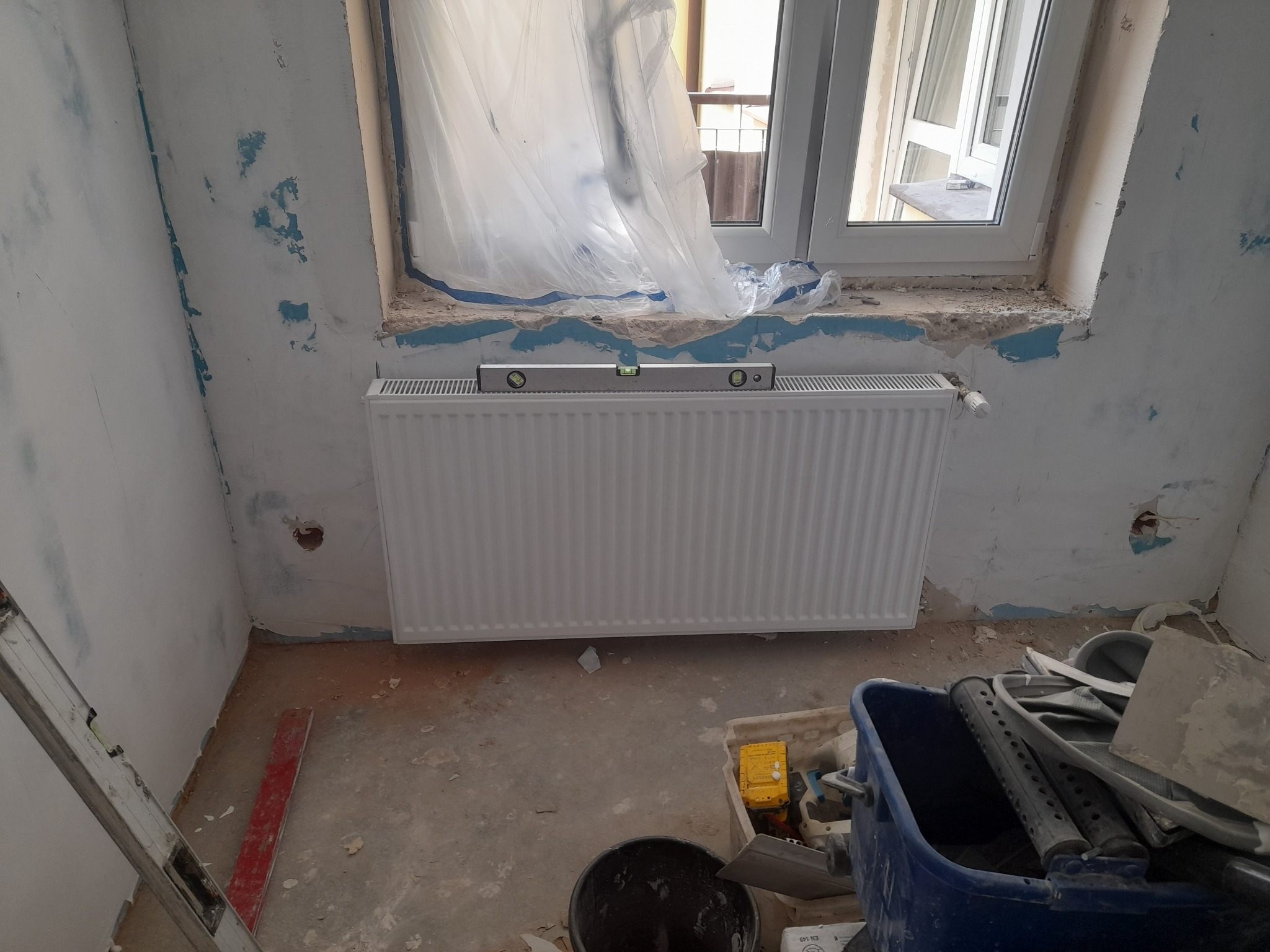 Montaż nowego grzejnika panelowego pod oknem, poziomica na górze, ściany w trakcie remontu, widoczne ślady po starej farbie i przygotowaniu do malowania.