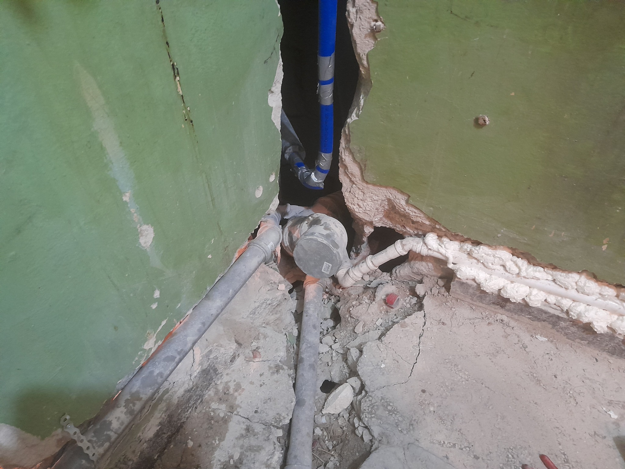 Fragment ściany z widocznymi starymi i nowymi rurami, niebieska rura owinięta taśmą wchodzi w otwór w ścianie, widoczne ślady po kuciu i pianka montażowa.