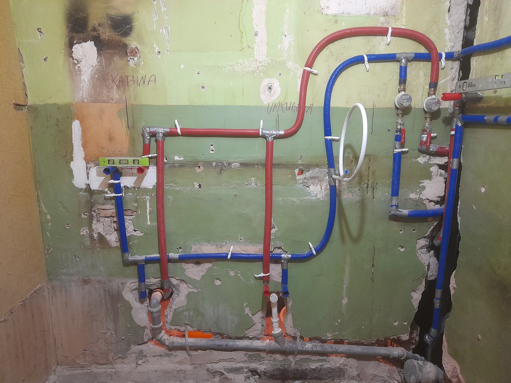 Instalacja wodna z rur PEX w kolorze czerwonym i niebieskim, częściowo zamontowana na starej, zielonej ścianie z widocznymi śladami po kuciu i napisem 'KABWA' oraz 'UMKIAGA'.