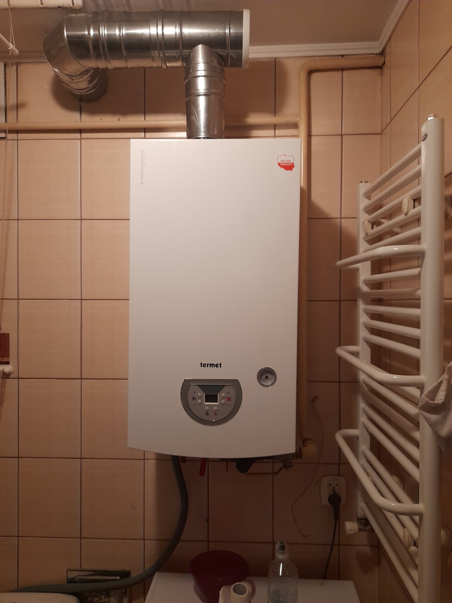 Biały piec gazowy marki Termet, z metalową rurą odprowadzającą spaliny, zamontowany na ścianie w łazience z beżowymi płytkami, obok grzejnika drabinkowego.