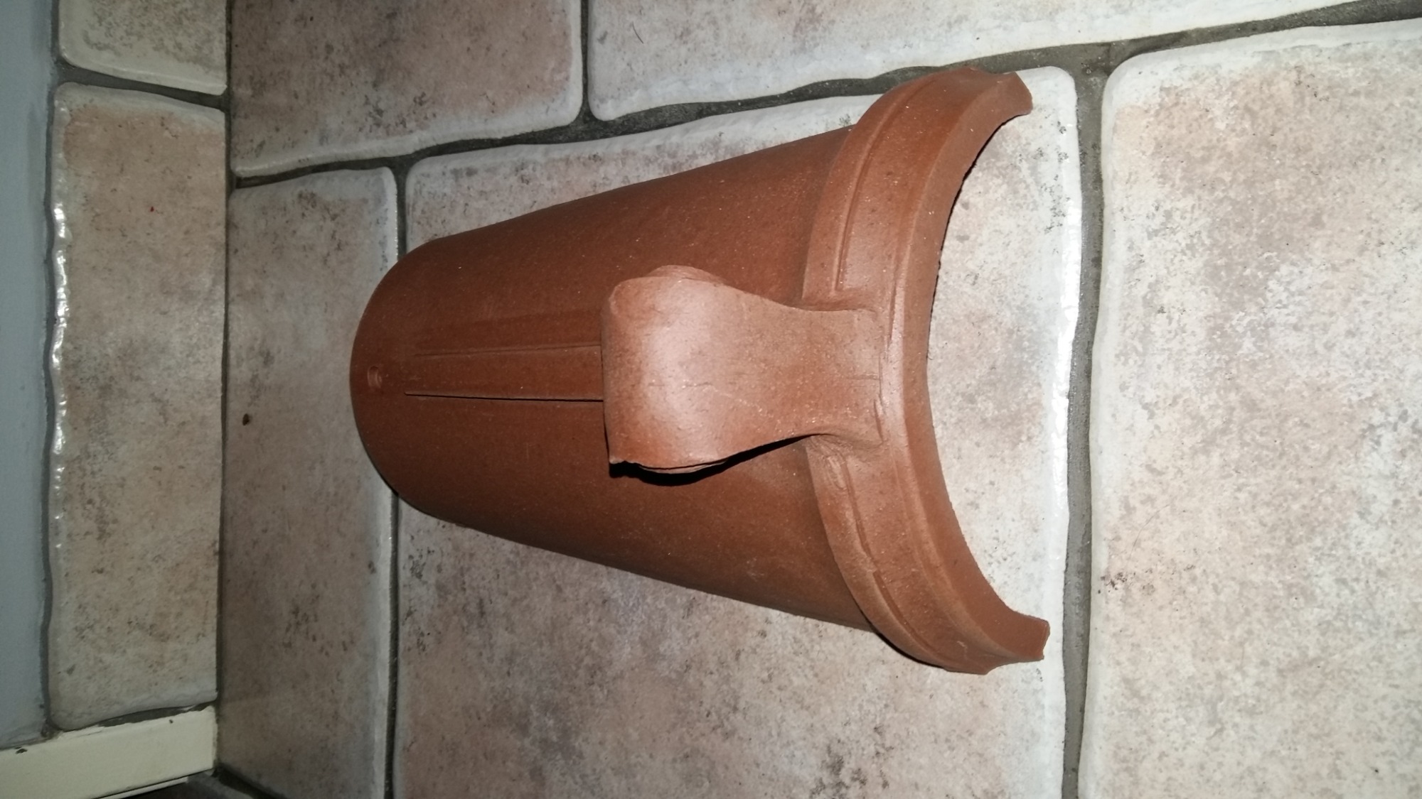 Brązowa kształtka ceramiczna do rur kanalizacyjnych z uchwytem, leżąca na podłodze wyłożonej płytkami imitującymi kamień.