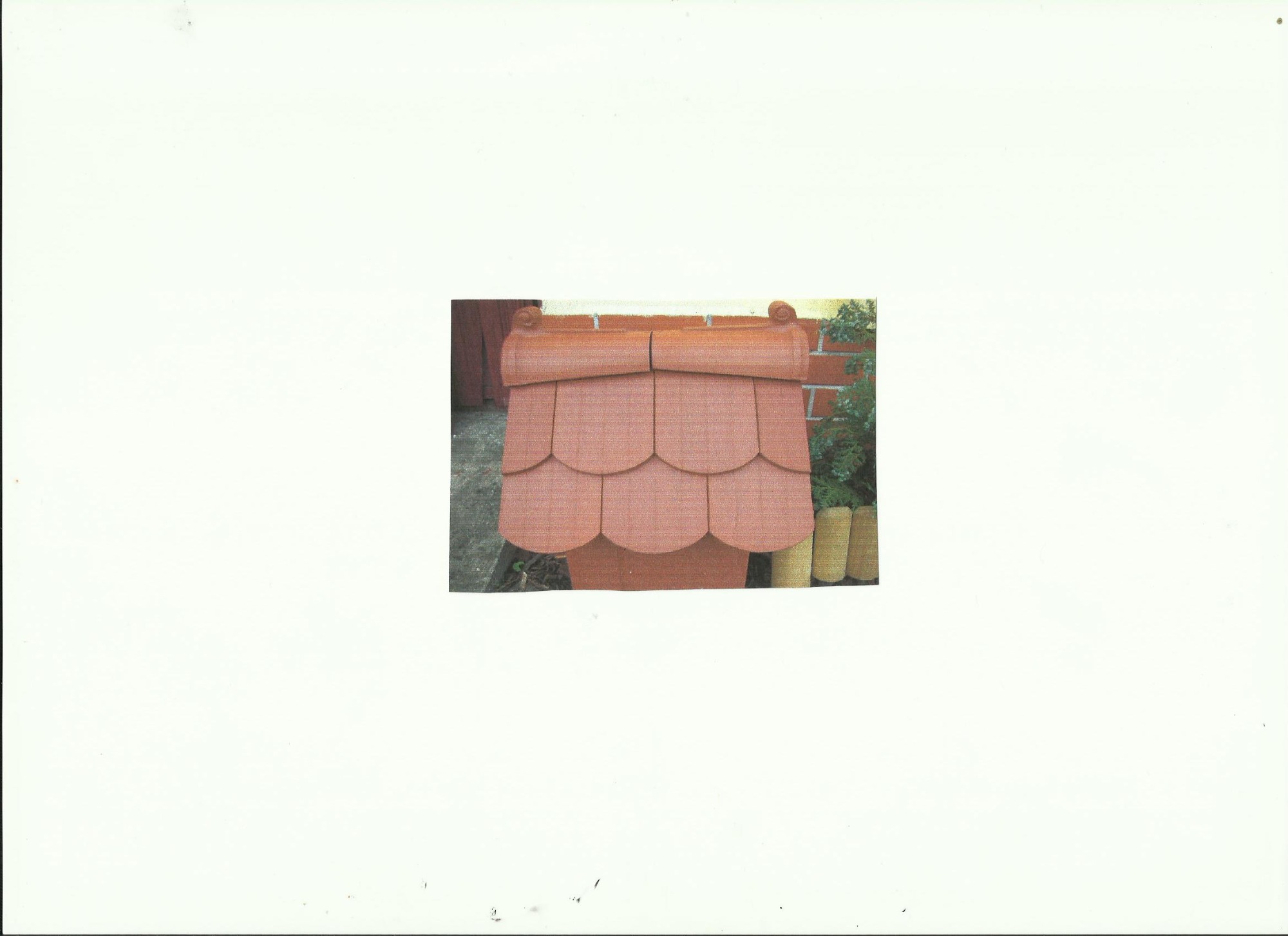 Model dachu z czerwonej dachówki ceramicznej w kształcie rybiej łuski, z dekoracyjnymi zakończeniami, prezentowany na tle ceglanej ściany i zieleni.