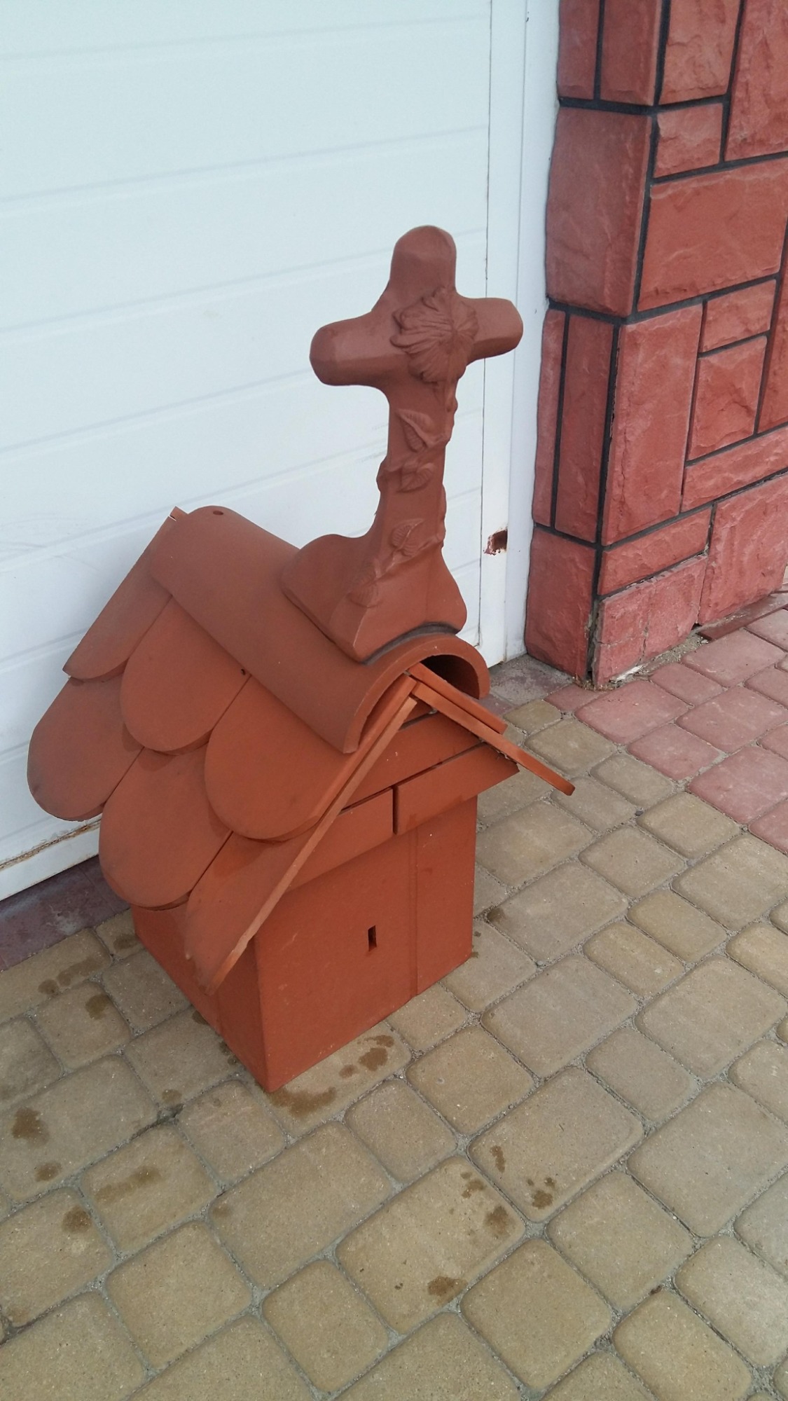 Ozdobna, ceramiczna skrzynka na listy w kształcie domku z dachem z dachówek i krzyżem z motywami roślinnymi na szczycie, ustawiona na kostce brukowej.